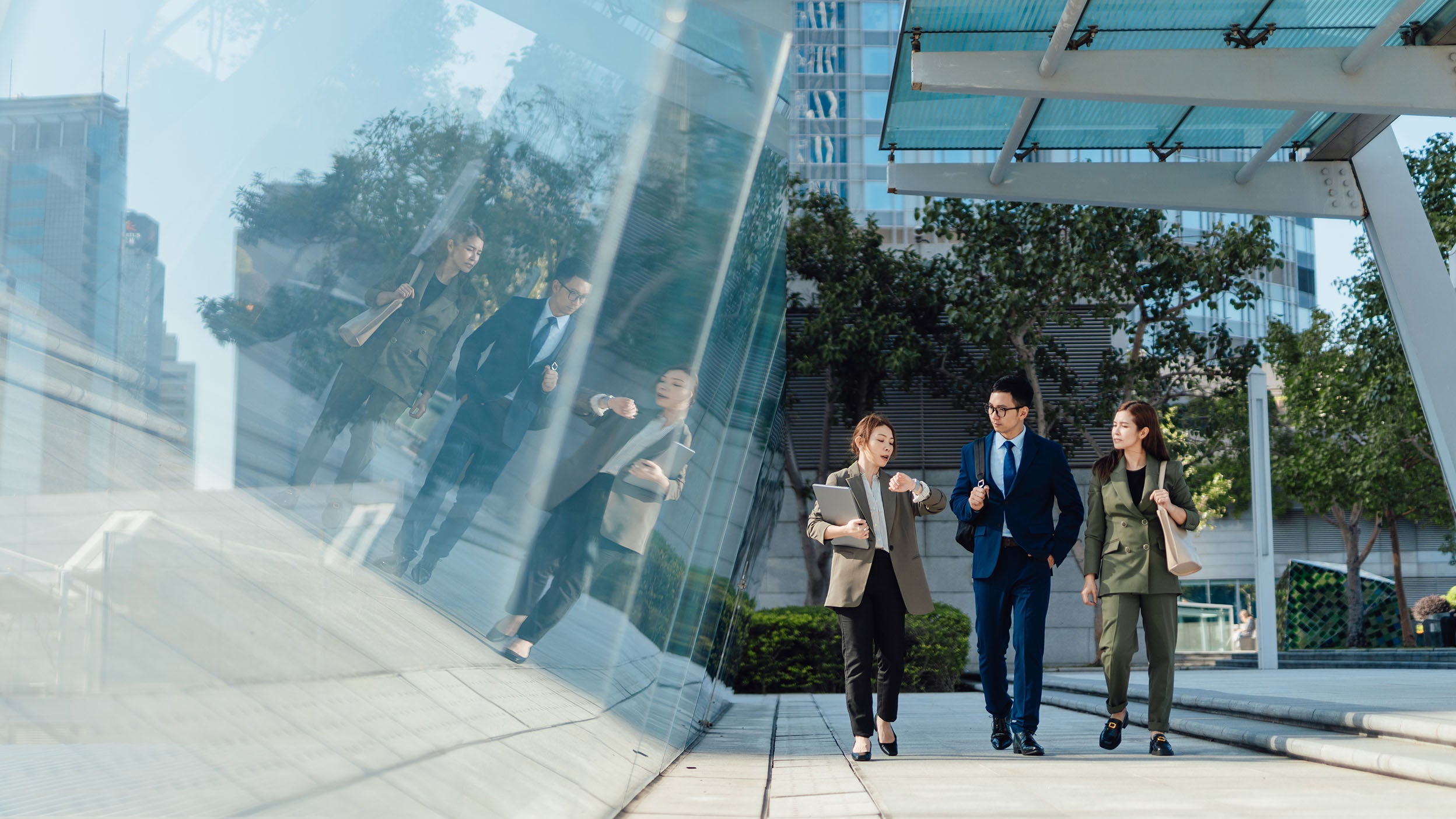 Business professionals walking together outside a modern office building, with their reflections visible in a curved glass facade.