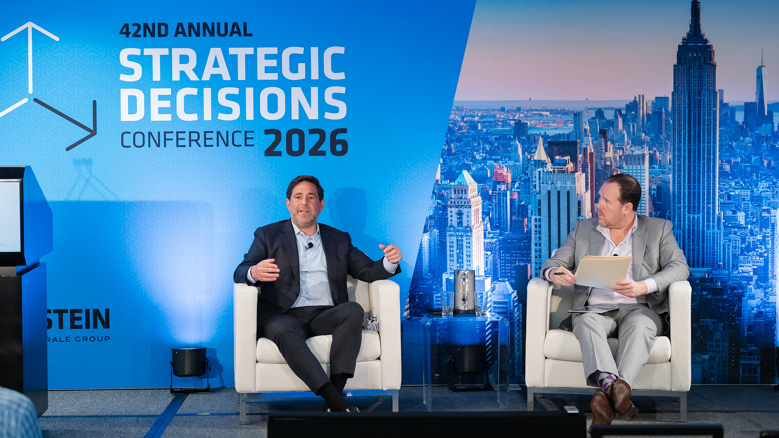 Two speakers seated on stage at the “42nd Annual Strategic Decisions Conference 2026,” with a blue branded backdrop and New York City skyline.
