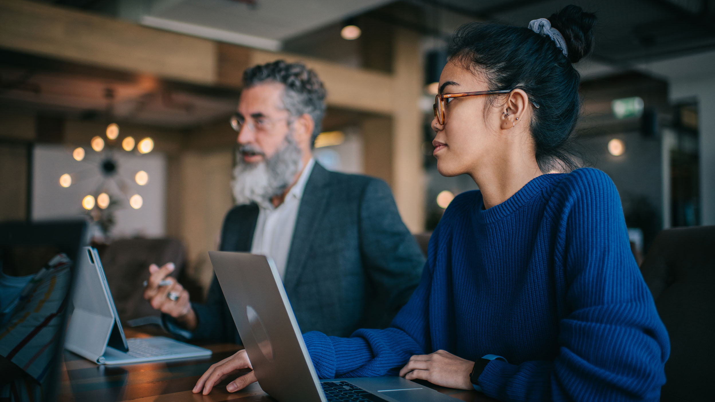 Young business woman in blue sweater, sitting next to elder businessman CEO while discussing work with team members across the desk in the office during afternoon.