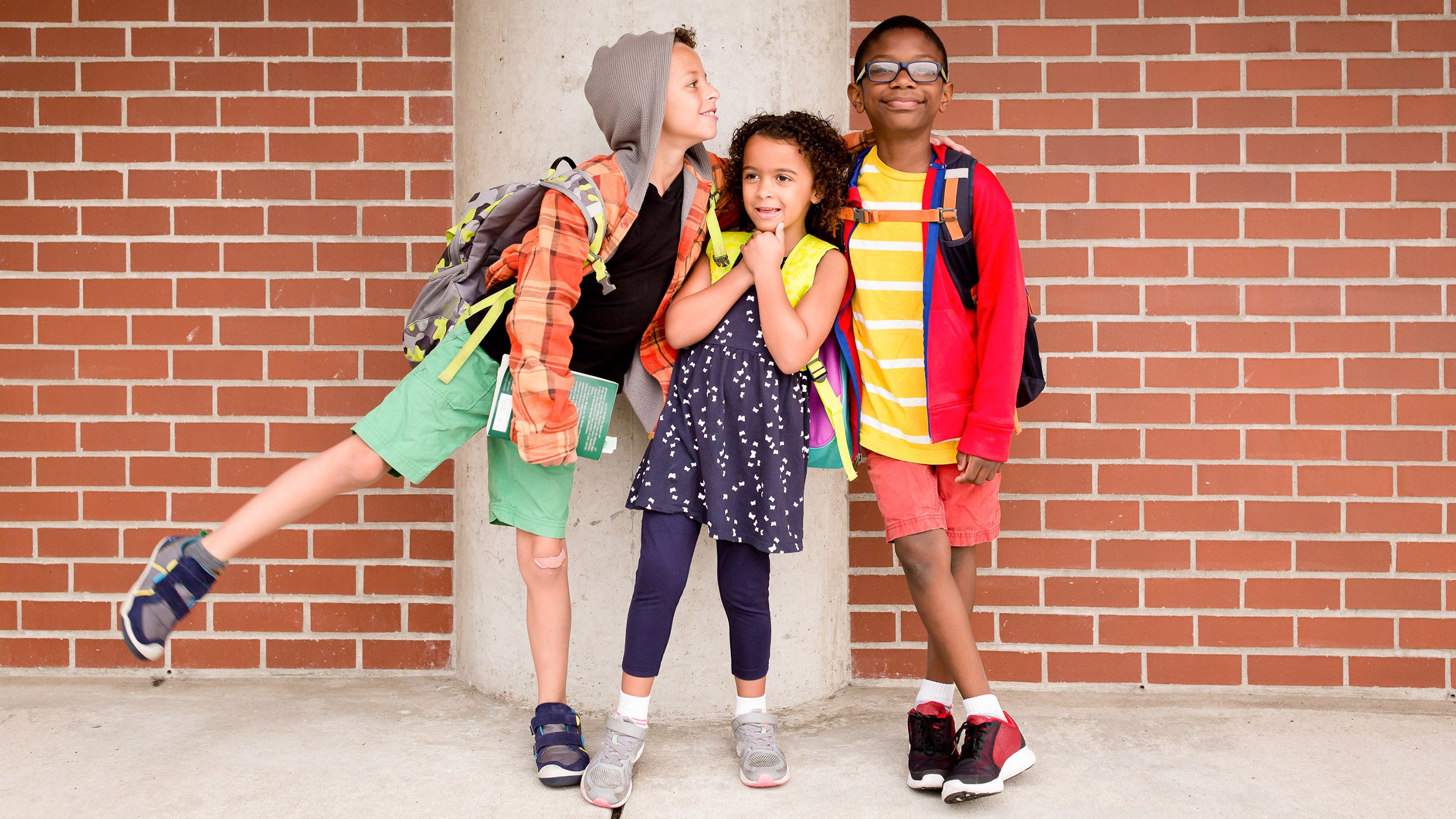 Children with backpacks standing together outside a school in front of a brick wall.