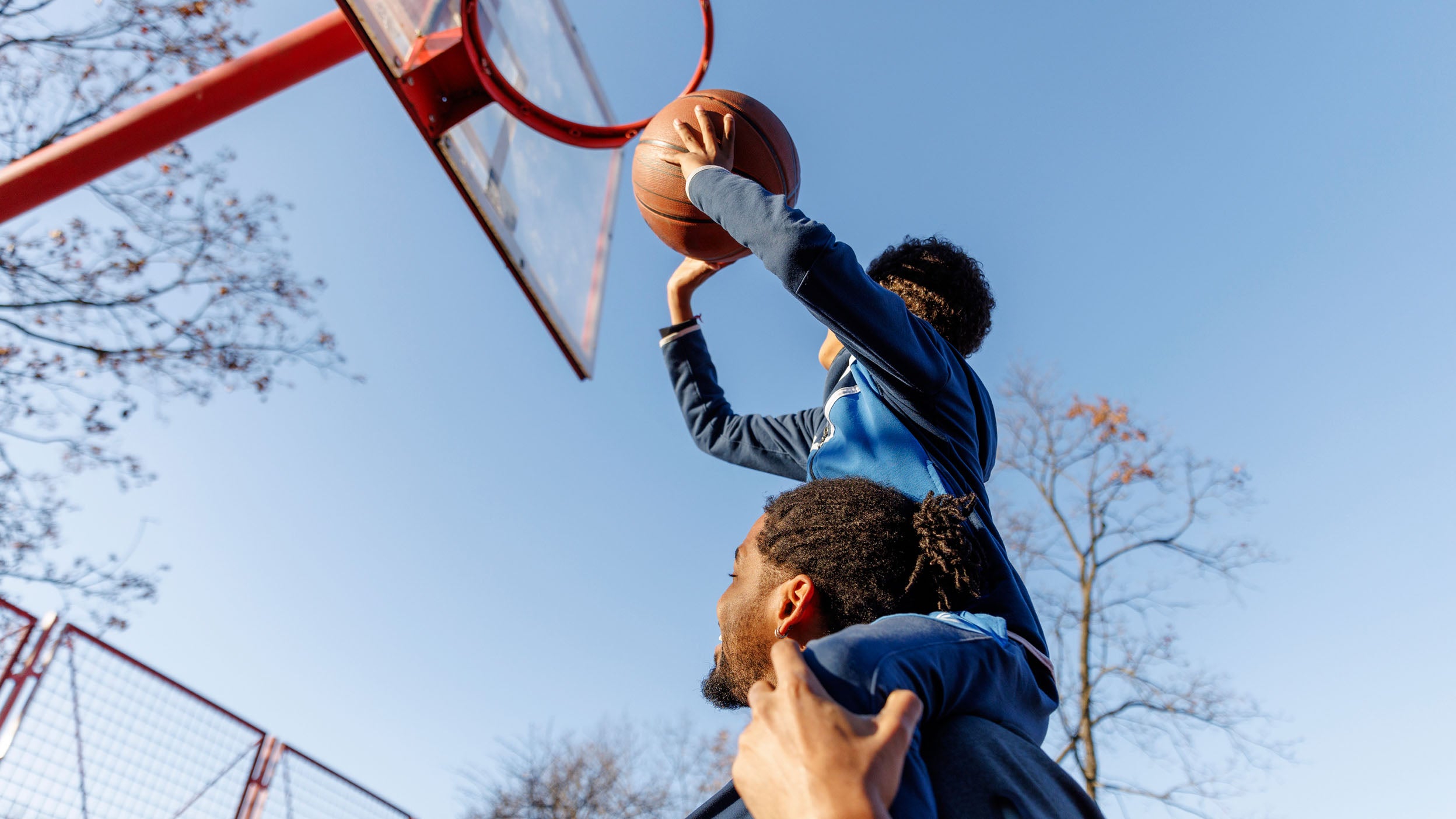 Low angle view of father carrying son on shoulders while assisting in scoring basket. Family is playing basketball on court against sky. They are enjoying competitive sports during sunny day.