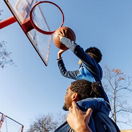 Low%20angle%20view%20of%20father%20carrying%20son%20on%20shoulders%20while%20assisting%20in%20scoring%20basket.%20Family%20is%20playing%20basketball%20on%20court%20against%20sky.%20They%20are%20enjoying%20competitive%20sports%20during%20sunny%20day.