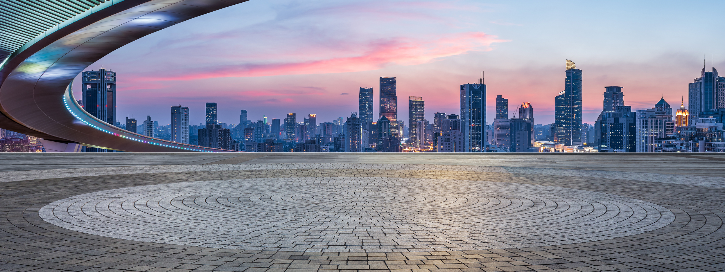 Modern city skyline at dusk viewed from a wide circular plaza, with a curved elevated walkway in the foreground and illuminated skyscrapers against a pink and blue sunset sky