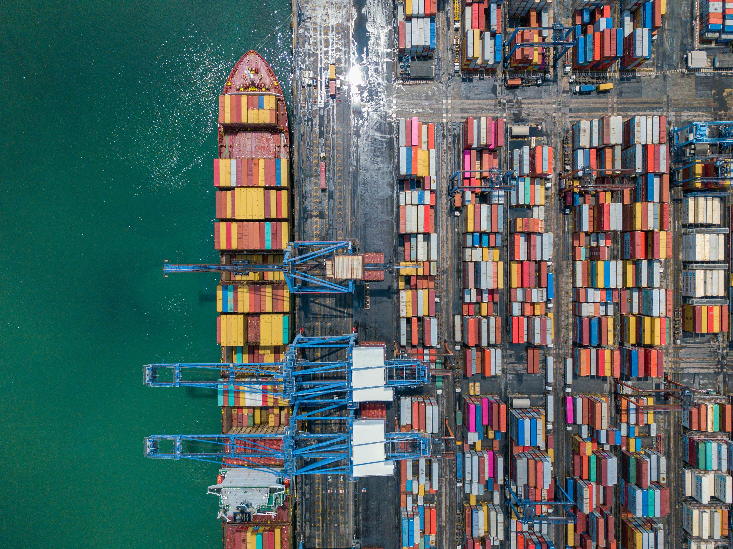 Aerial view of a container ship at a busy port with colorful shipping containers and blue cranes.