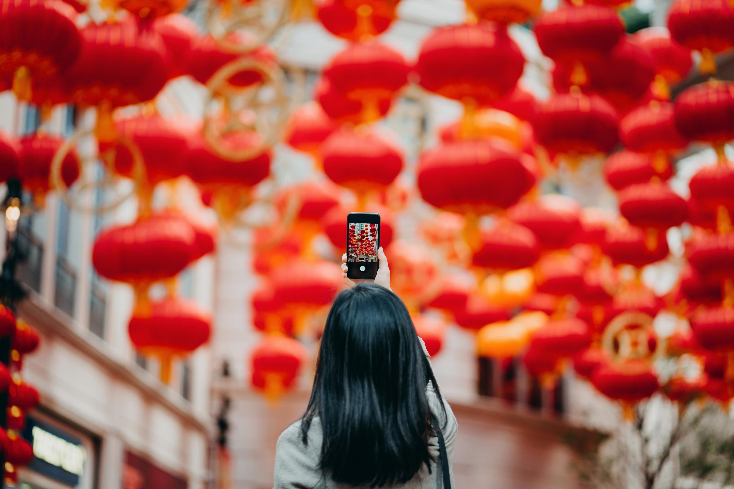 Rear view of woman taking photos of traditional Chinese red lanterns with smartphone on city street