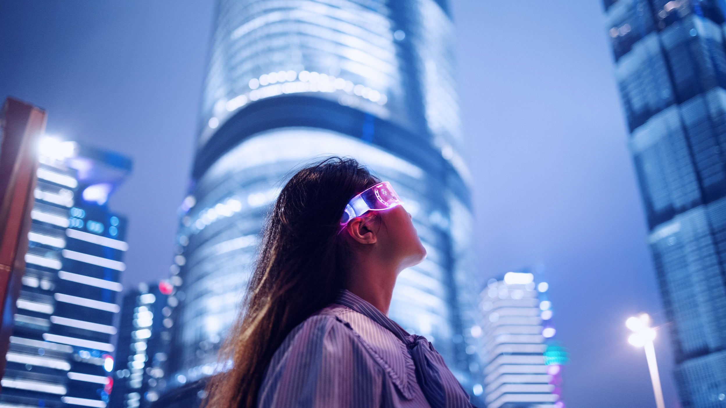 Person in glowing glasses looking up at a lit skyscraper in a nighttime cityscape.