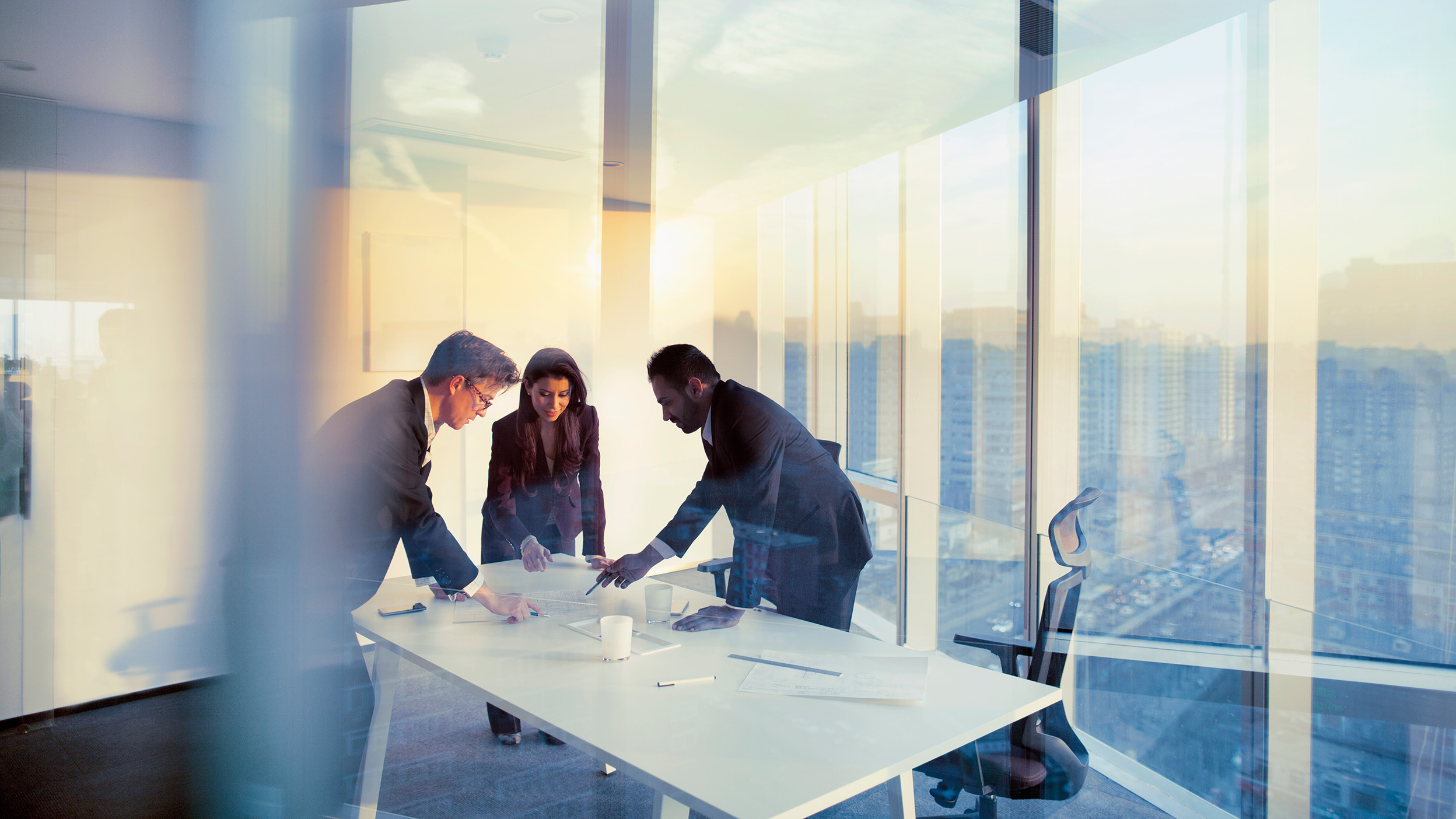 Three professionals reviewing documents together at a table in a bright office with floor-to-ceiling windows and city views.