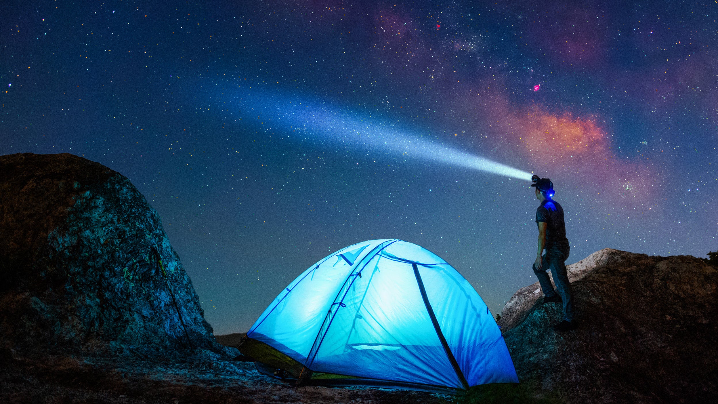 A person wearing a headlamp stands beside a glowing blue tent between rocks, looking up at a starry night sky.