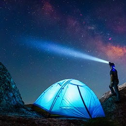 A%20person%20wearing%20a%20headlamp%20stands%20beside%20a%20glowing%20blue%20tent%20between%20rocks,%20looking%20up%20at%20a%20starry%20night%20sky.