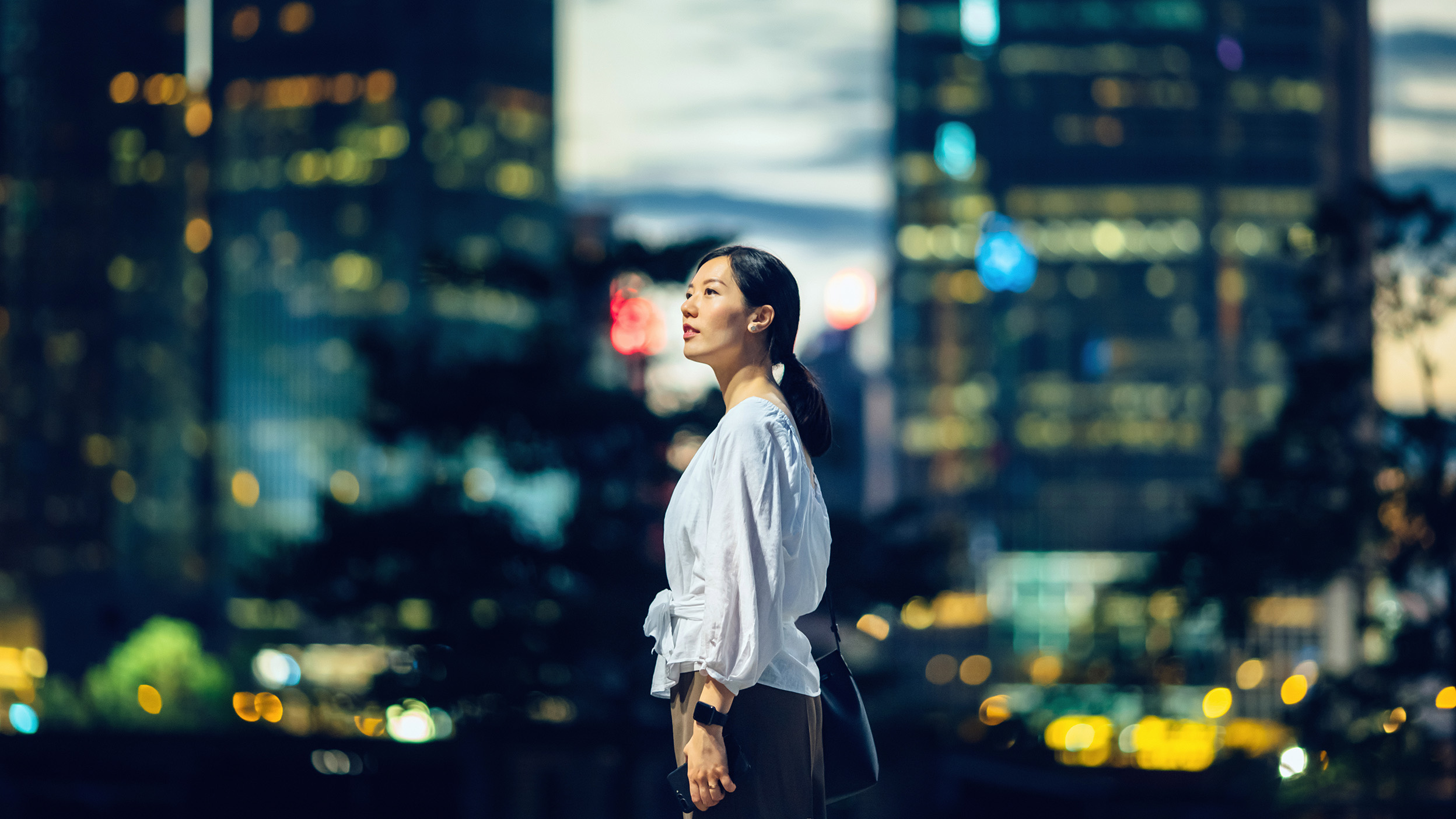 Confident young Asian woman standing against illuminated city buildings and thinking of her career path.