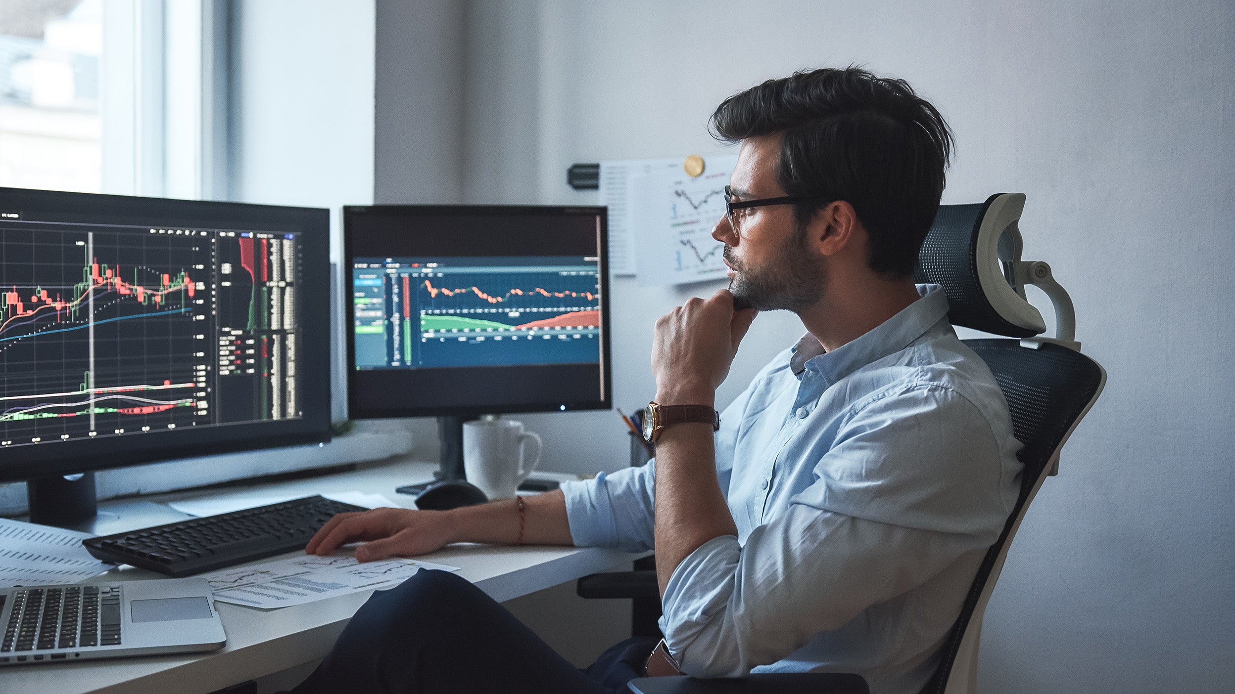Side view of successful trader or businessman in formal wear and eyeglasses working with charts and market reports on computer screens in his modern office.