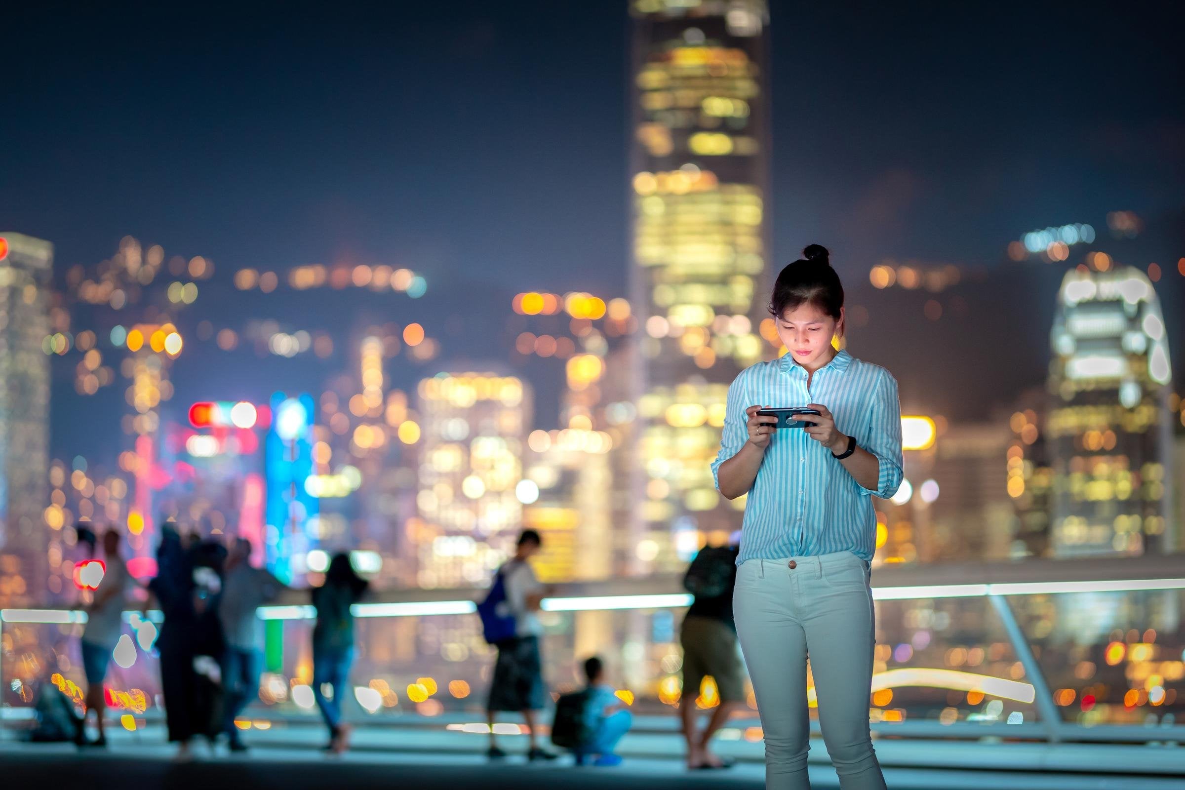 Asian woman using smartphone at night with Victoria Harbor in background.