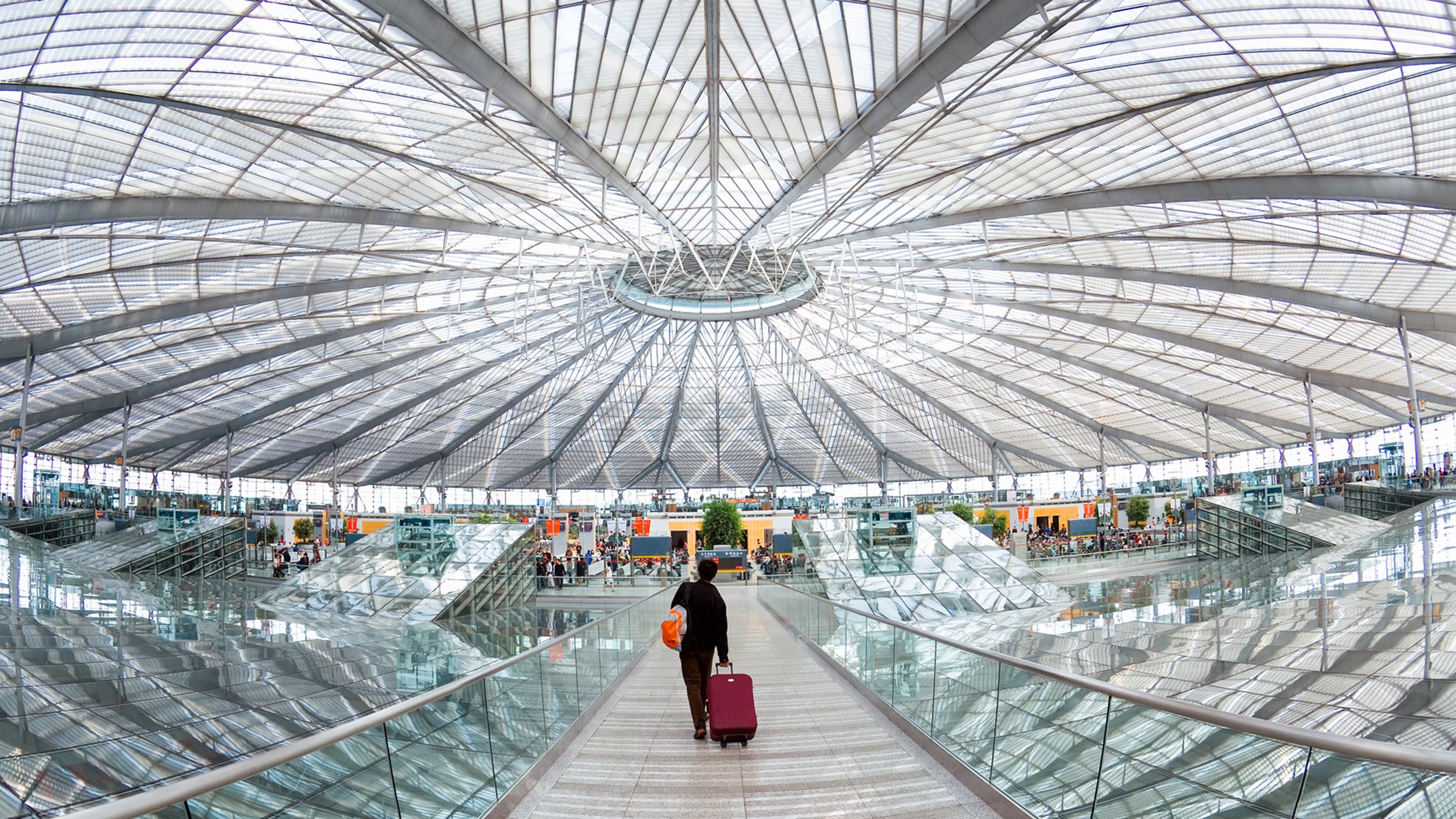 A traveller walking along a glass walkway inside a large, modern airport terminal with a bright, dome‑shaped ceiling.