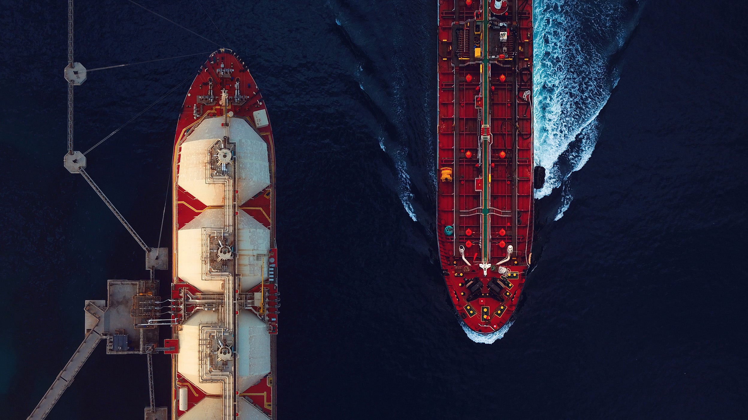 Bird’s-eye view of two passing ships on deep blue water, one solid red and the other red and white. Waves ripple beneath the red ship, creating a contrast with the calm sea.