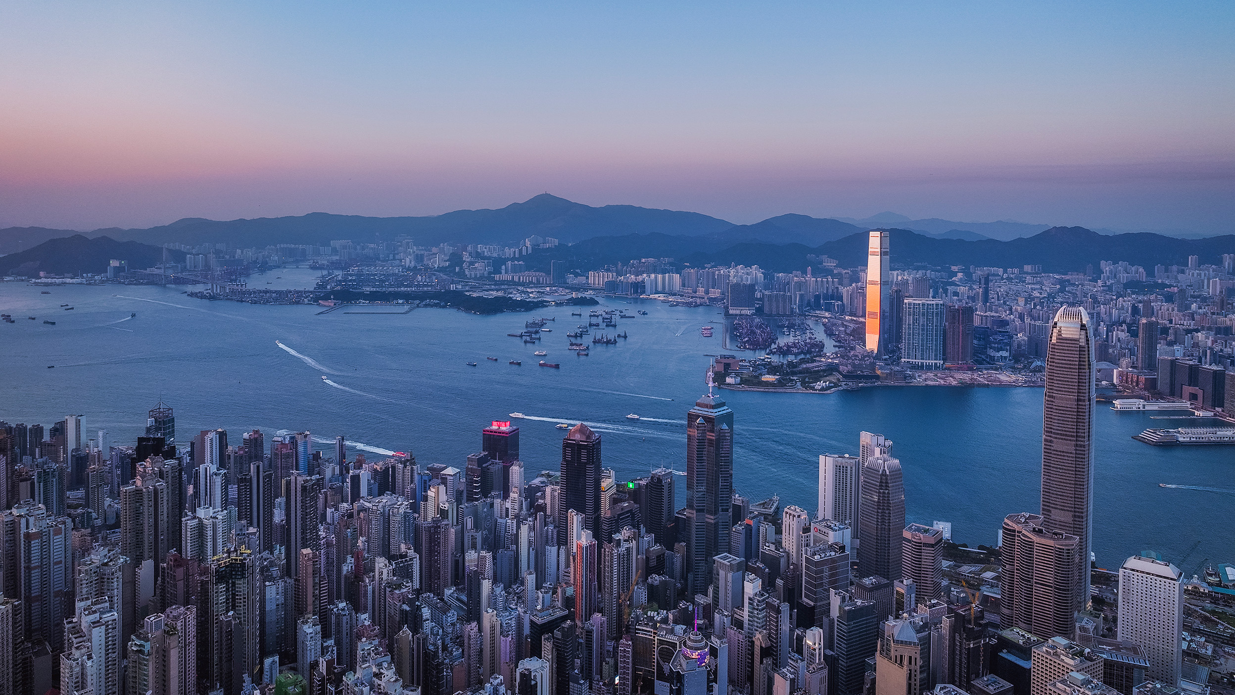 Hong Kong skyline at dusk overlooking Victoria Harbour, with dense high-rise buildings in the foreground, illuminated skyscrapers along the waterfront, and boats moving across the water against a backdrop of mountains.