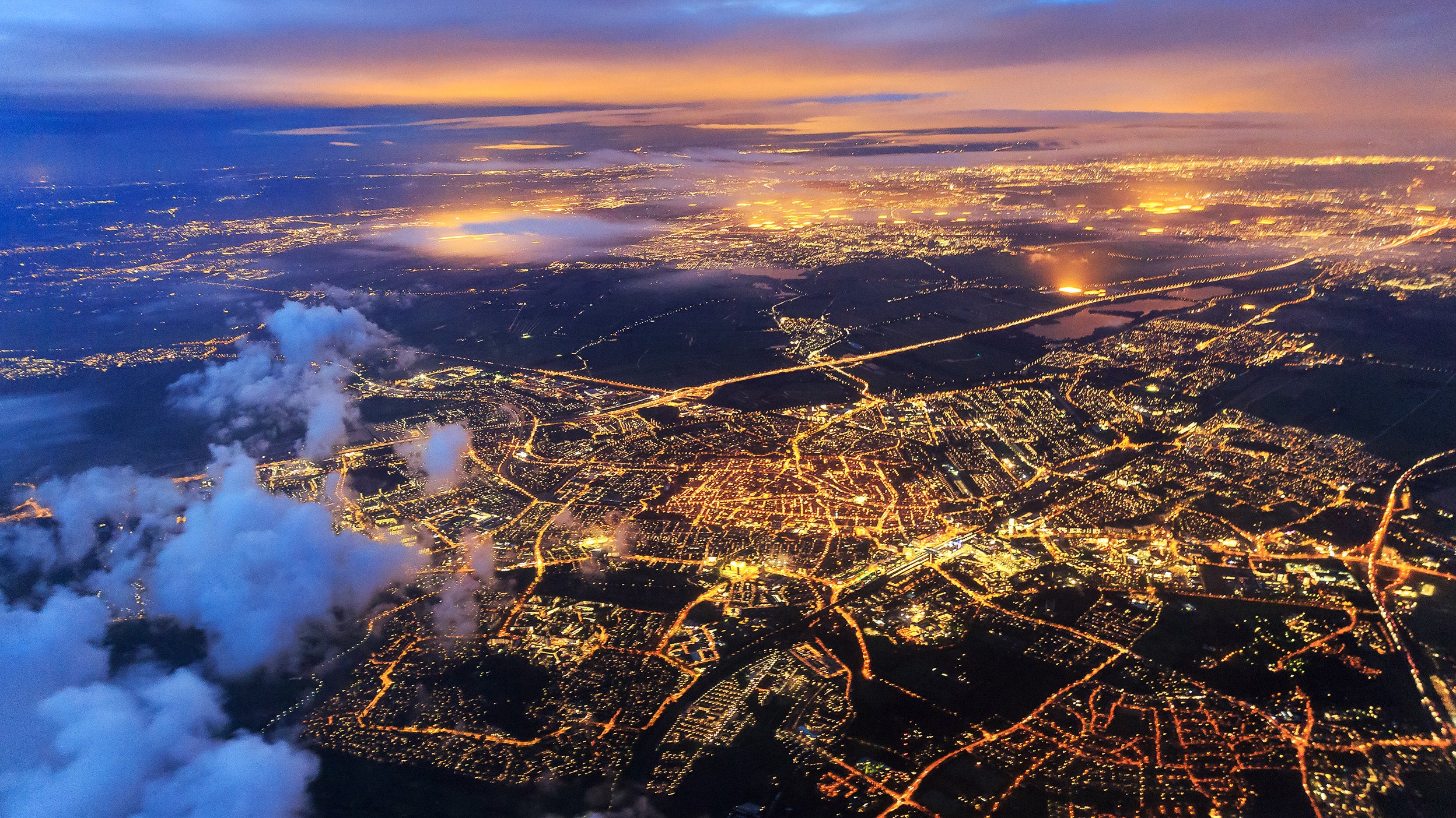 Aerial view of a city at dusk, with illuminated road networks and neighbourhoods glowing against low clouds.