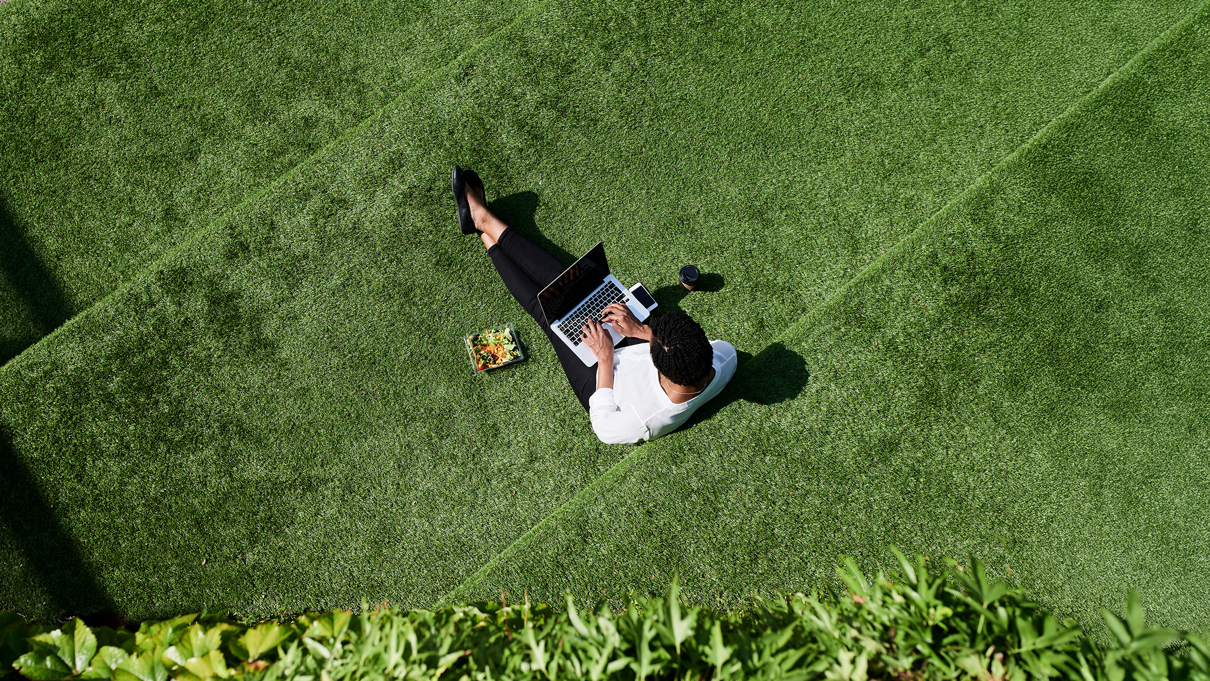 Person using a laptop on grass with coffee and lunch nearby