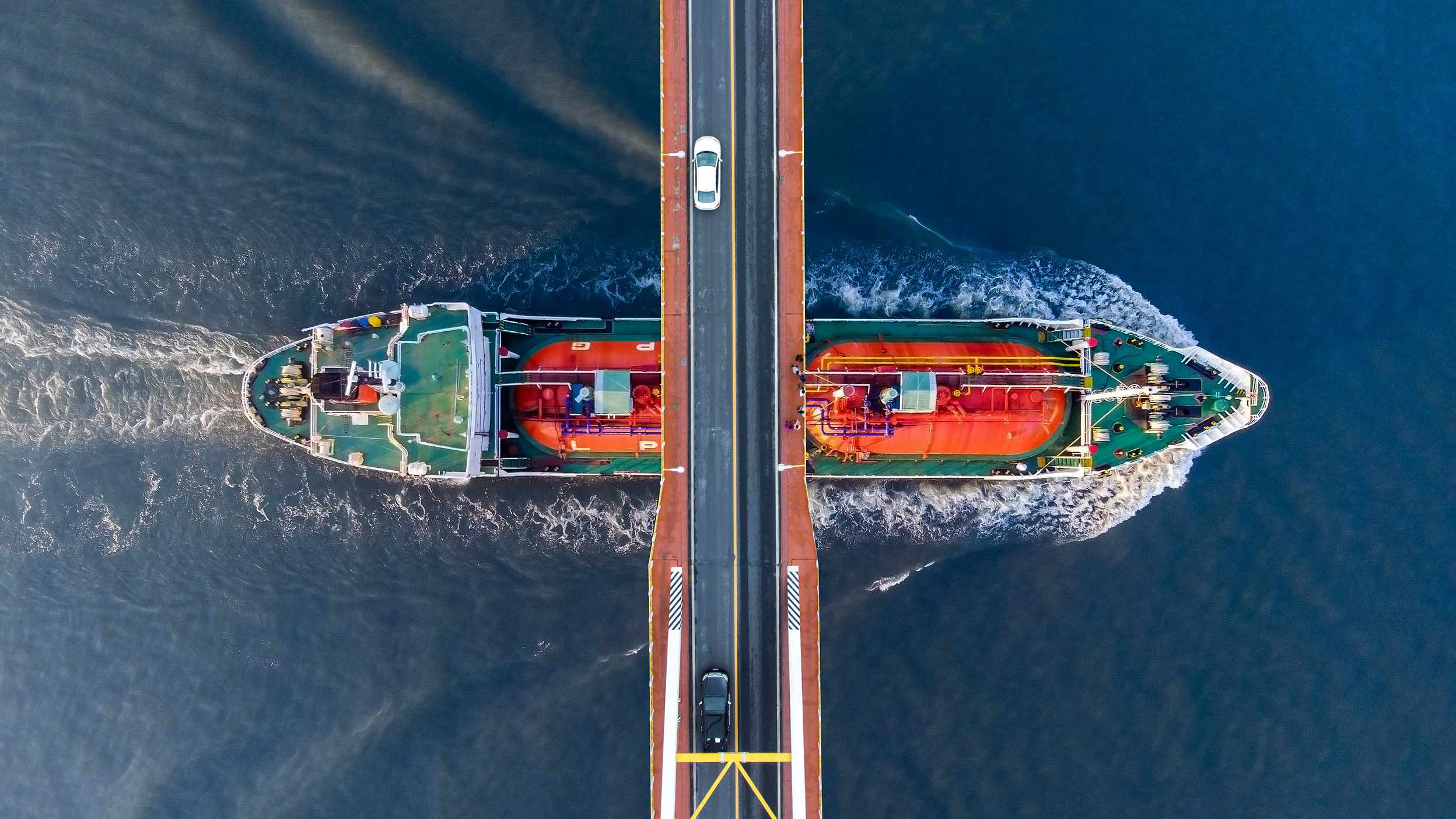 Overhead view of two cargo ships passing beneath a bridge, with vehicles crossing above and wakes visible in the surrounding water.
