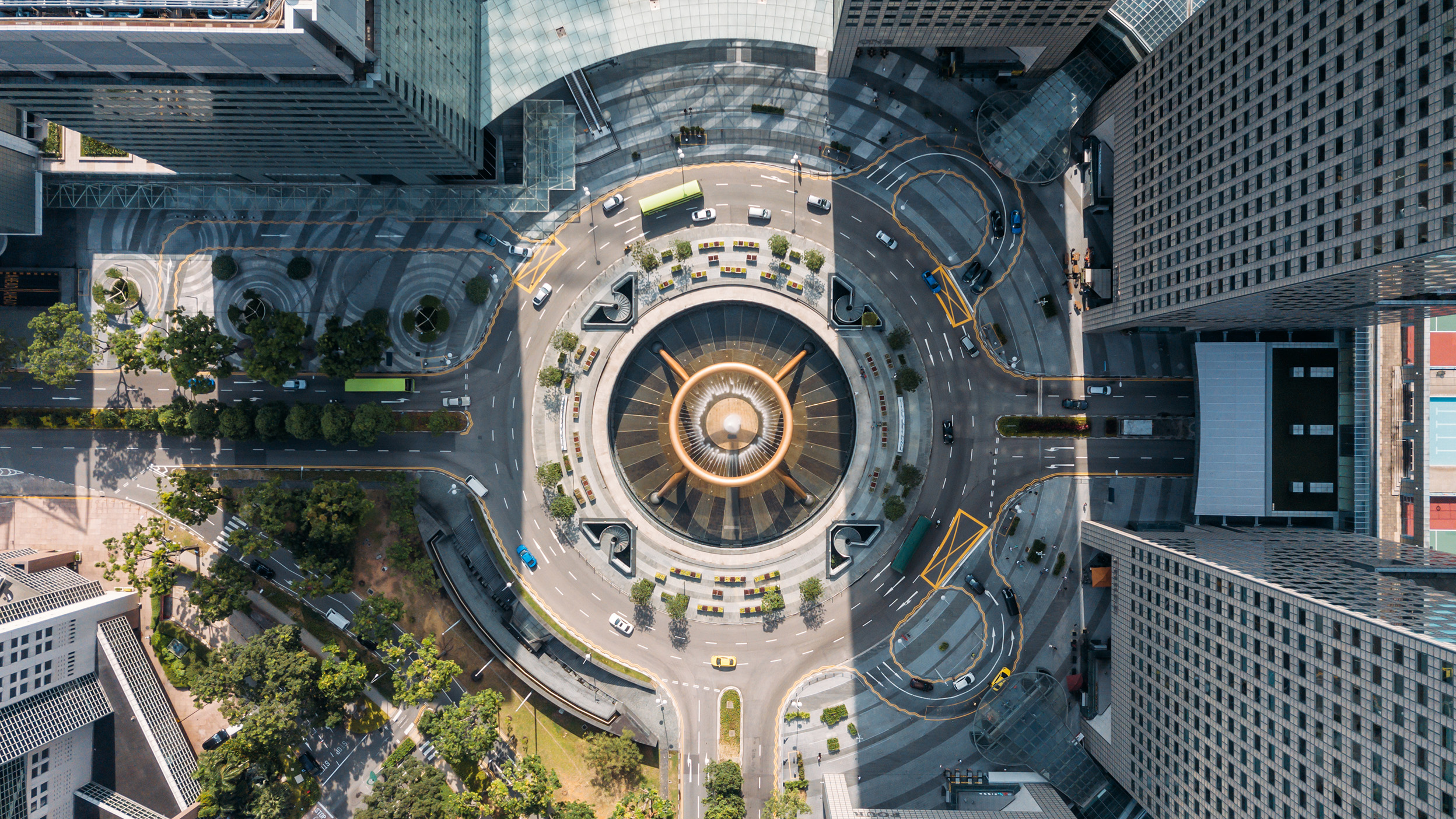 Top-down aerial view of a large circular roundabout with a central fountain, encircled by multi-lane roads, cars, greenery, and modern high-rise buildings.