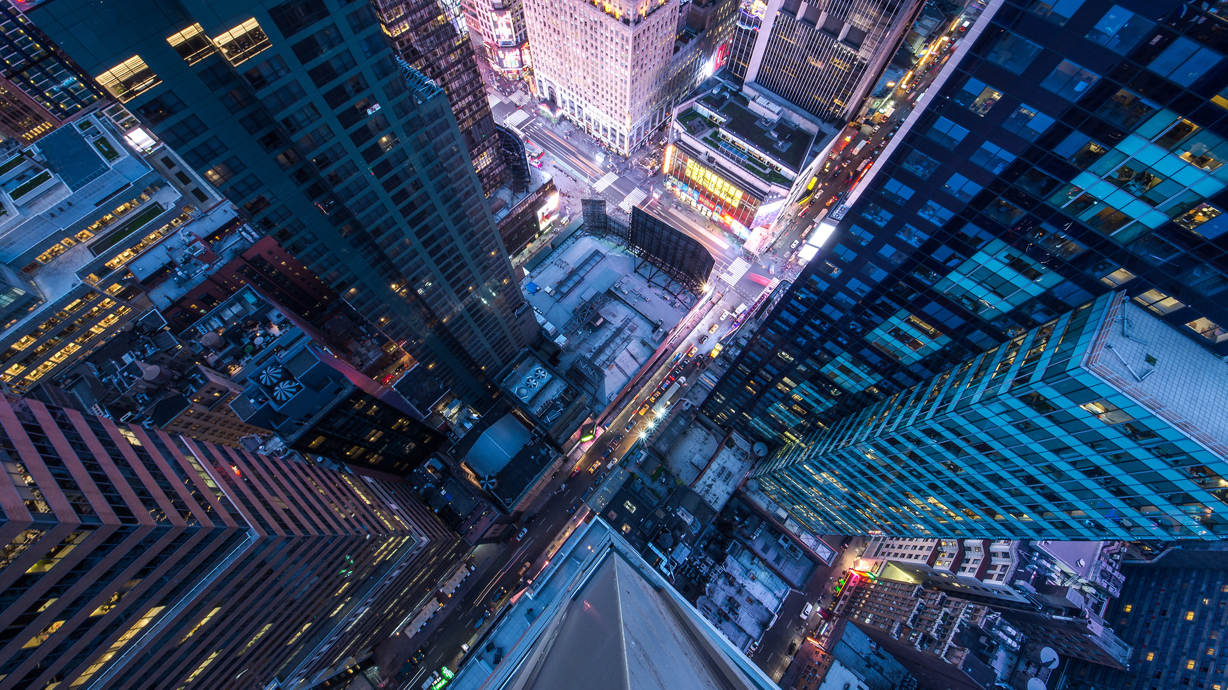 Aerial view looking straight down between tall city skyscrapers, with intersecting streets below illuminated by headlights and building lights at dusk.