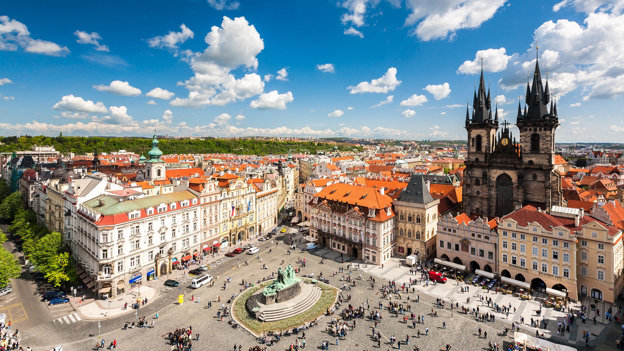 Aerial view of Prague's Old Town Square with colourful historic buildings, a central statue, and the Church of Our Lady before Týn under a partly cloudy sky.