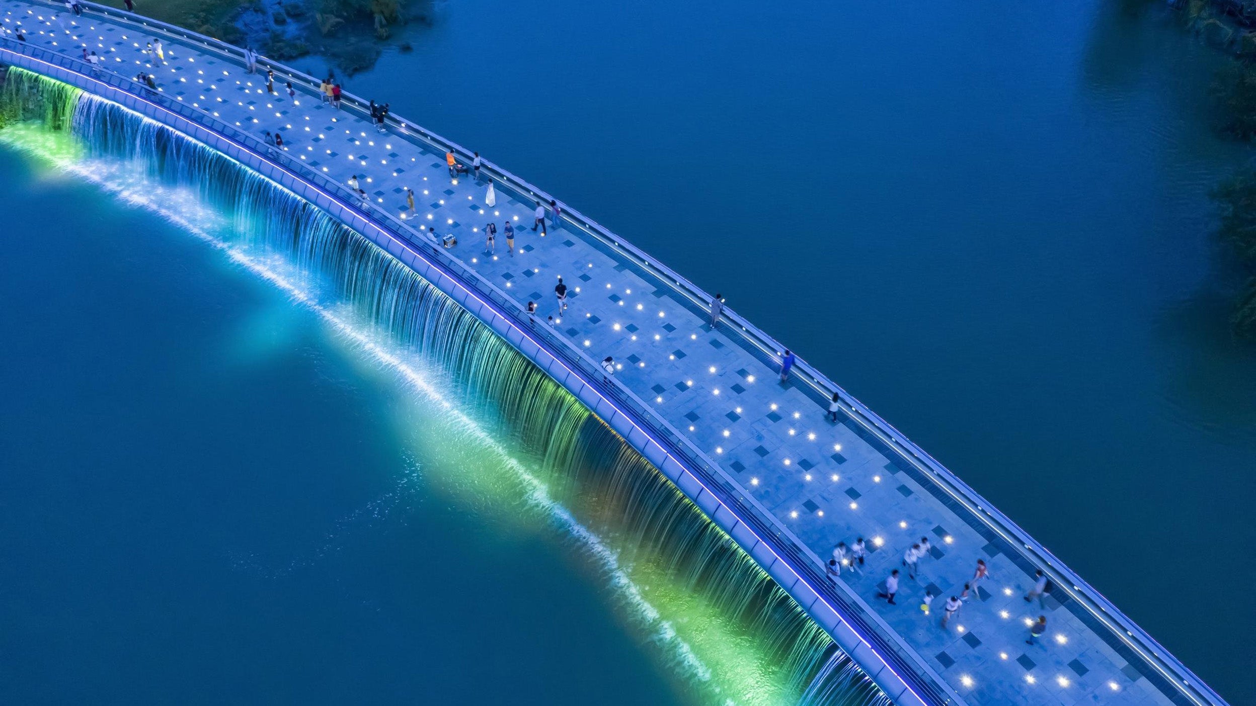 Aerial view of a modern pedestrian bridge illuminated with bright white lights, spanning across a calm blue river. Several people are walking along the bridge, enjoying the scenic and artistic design. 