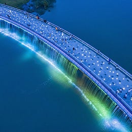 Aerial%20view%20of%20a%20modern%20pedestrian%20bridge%20illuminated%20with%20bright%20white%20lights,%20spanning%20across%20a%20calm%20blue%20river.%20Several%20people%20are%20walking%20along%20the%20bridge,%20enjoying%20the%20scenic%20and%20artistic%20design.%20