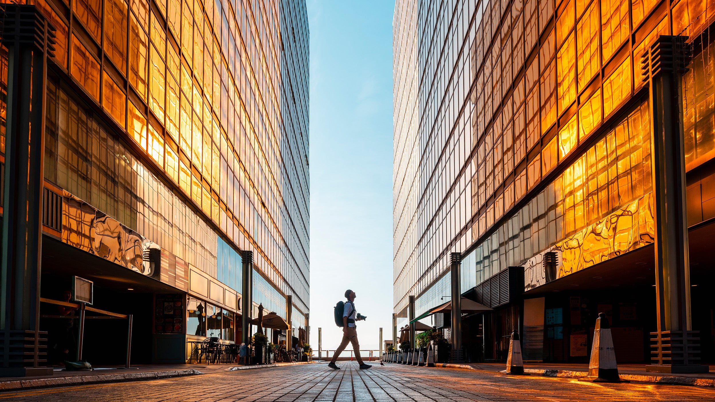 Person with backpack walking between tall glass buildings at sunrise or sunset.