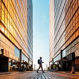 Person%20with%20backpack%20walking%20between%20tall%20glass%20buildings%20at%20sunrise%20or%20sunset.%20