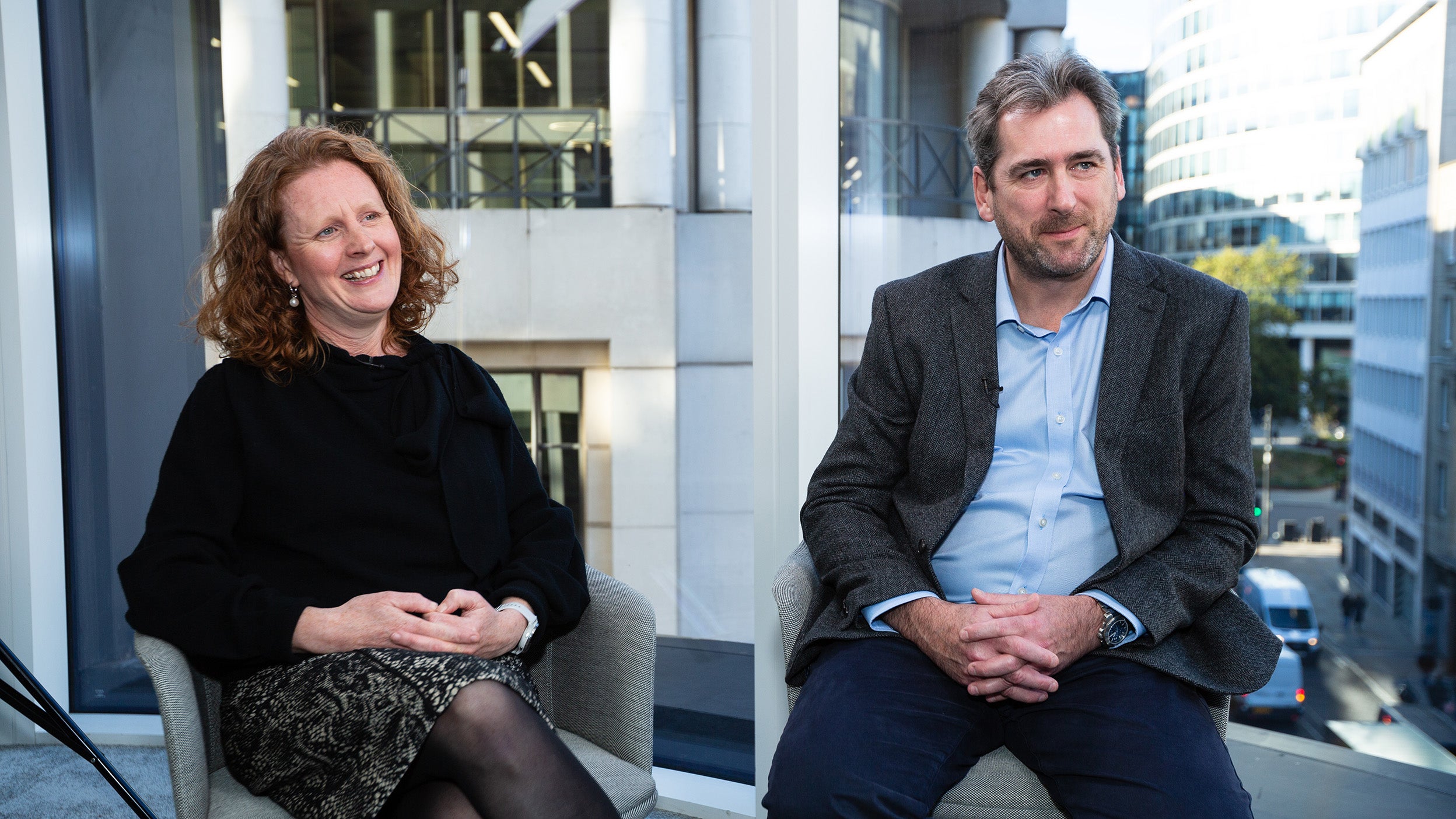 Two people seated indoors near large windows with city buildings visible outside.