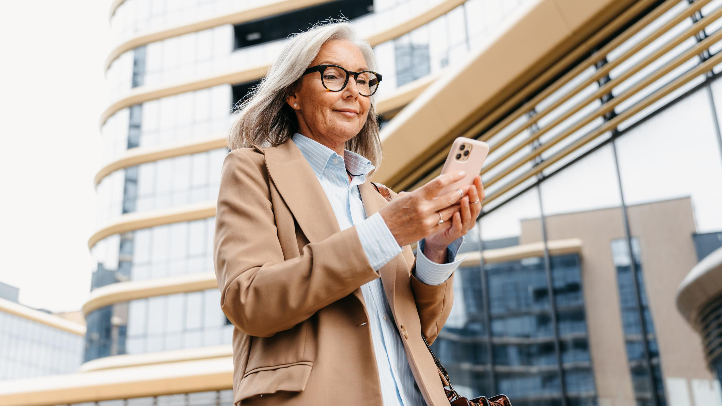A woman with grey hair in business clothes smiles while using a mobile phone standing outdoors in the business center of a modern city
