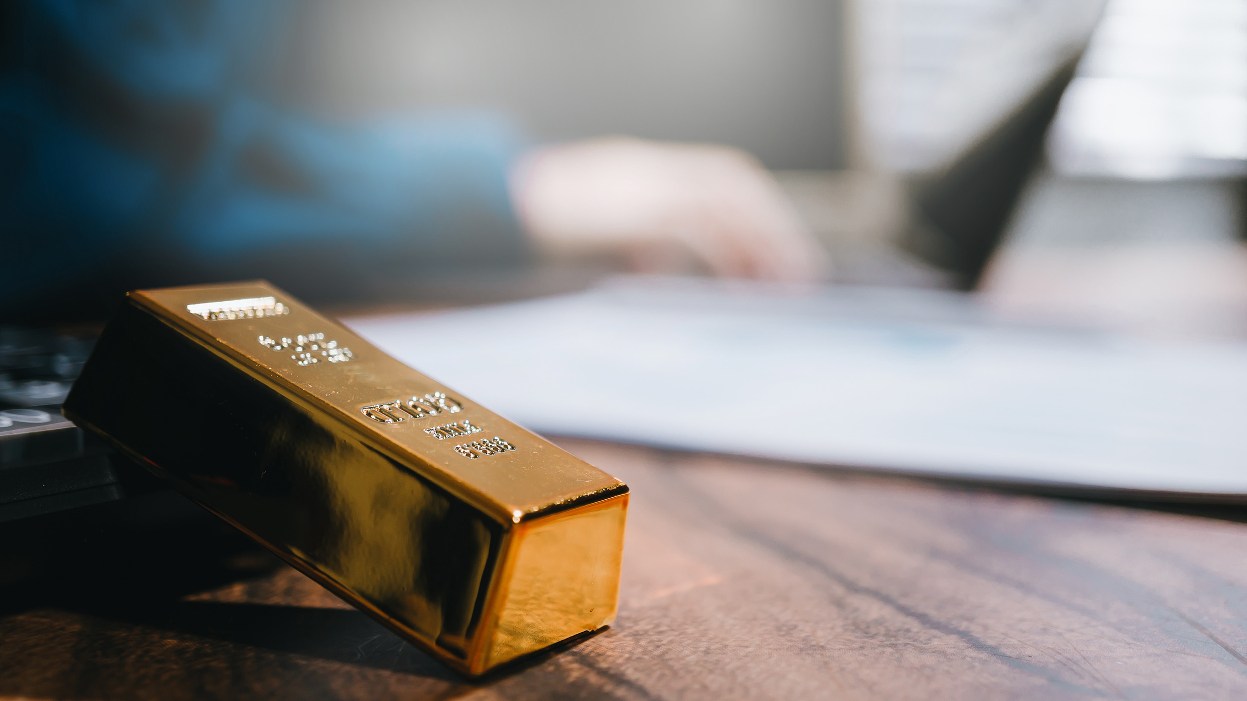 Gold bar resting on a wooden desk in an office setting, with documents and a laptop blurred in the background.