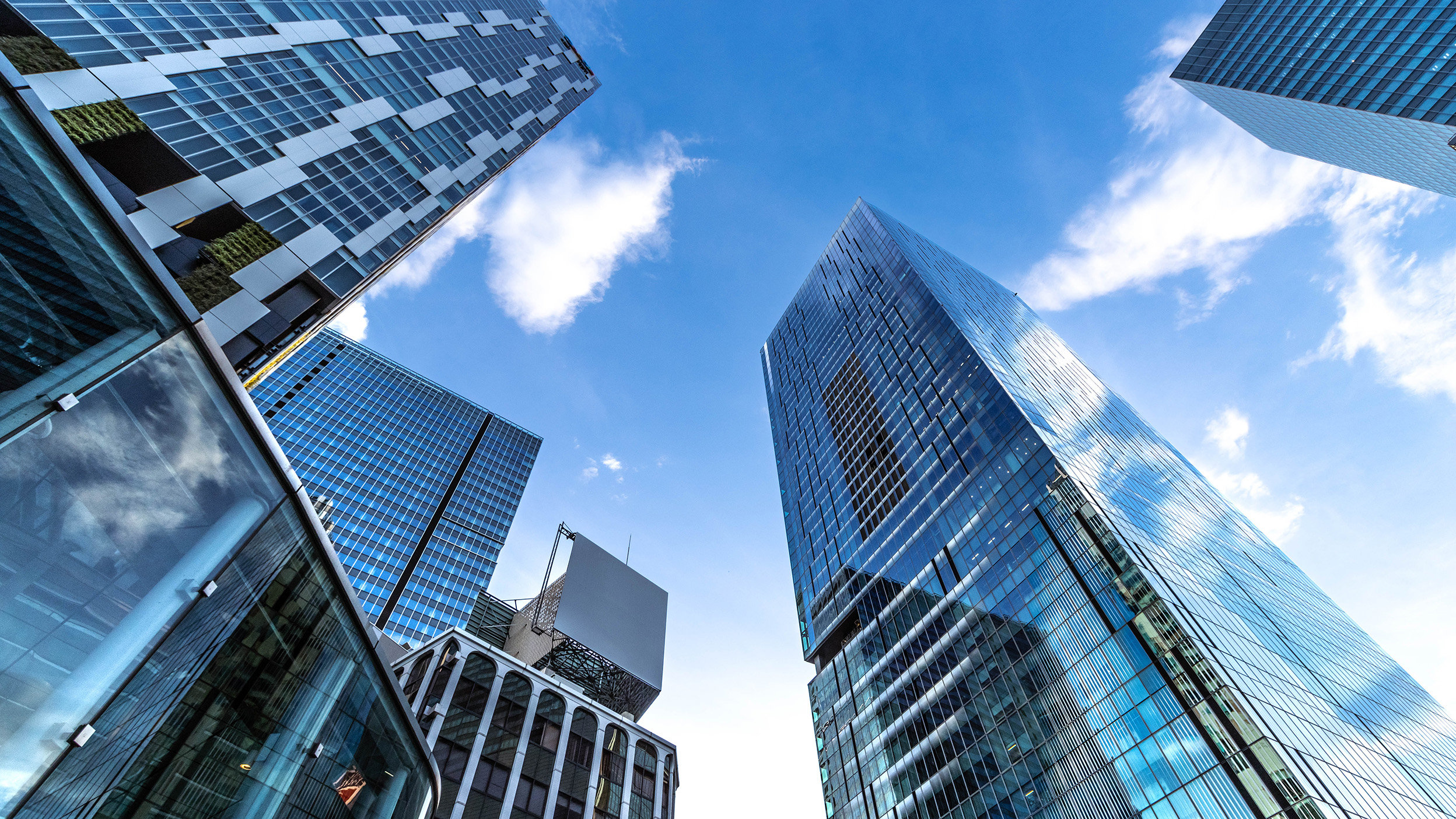 Looking up at modern glass office skyscrapers converging toward a bright blue sky, symbolising scale, structure, and forward-looking institutional investment.
