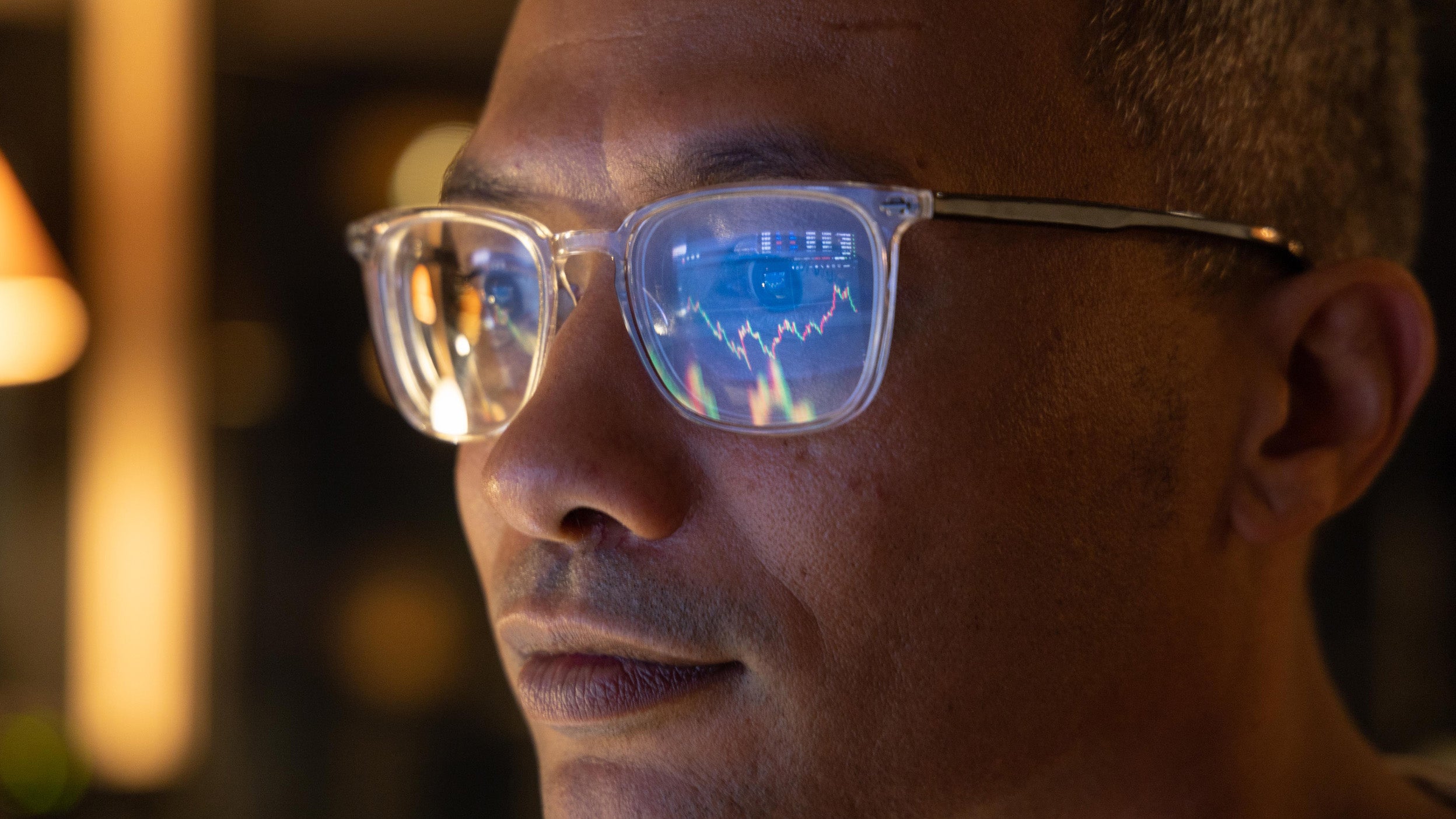 Close of man wearing glasses staring at the screen; the screen is reflected in his glasses showing he's looking at charts