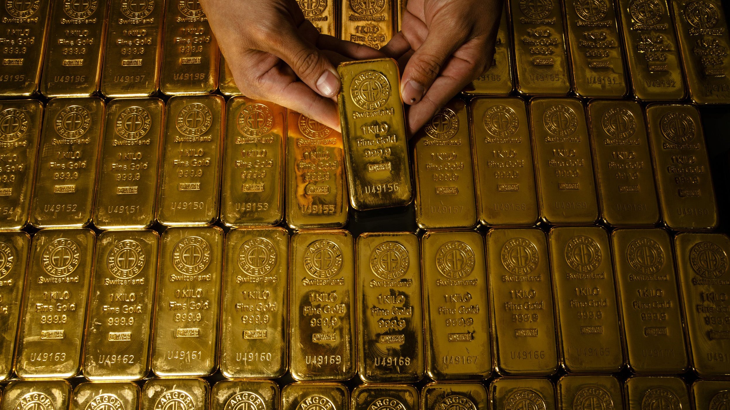 A person holding a gold bar among rows of gold bars, each marked with '1000g' and '999.9'.
