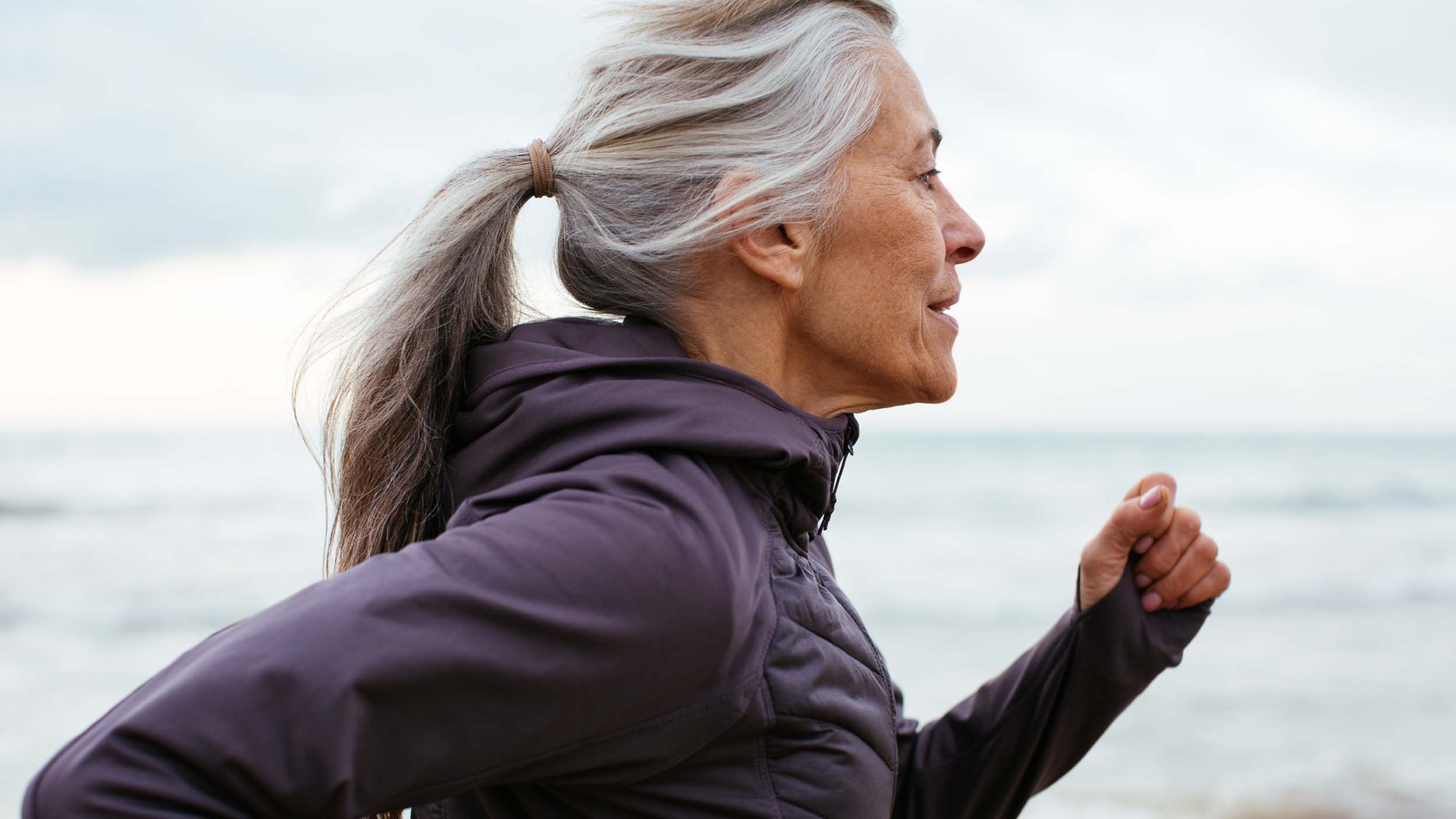 Persona anziana con capelli legati in una coda di cavallo, che indossa una giacca scura, che fa jogging vicino al mare con un cielo nuvoloso sullo sfondo.