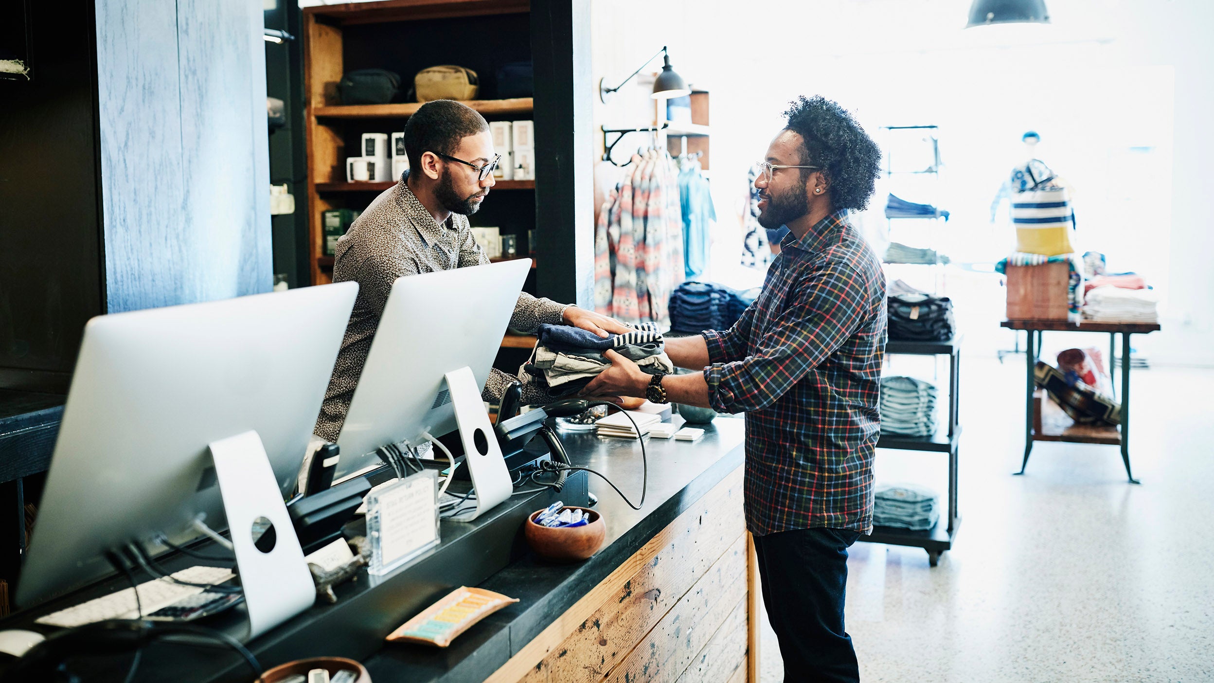 Man handing clothes to shopkeeper at register in men’s clothing boutique.