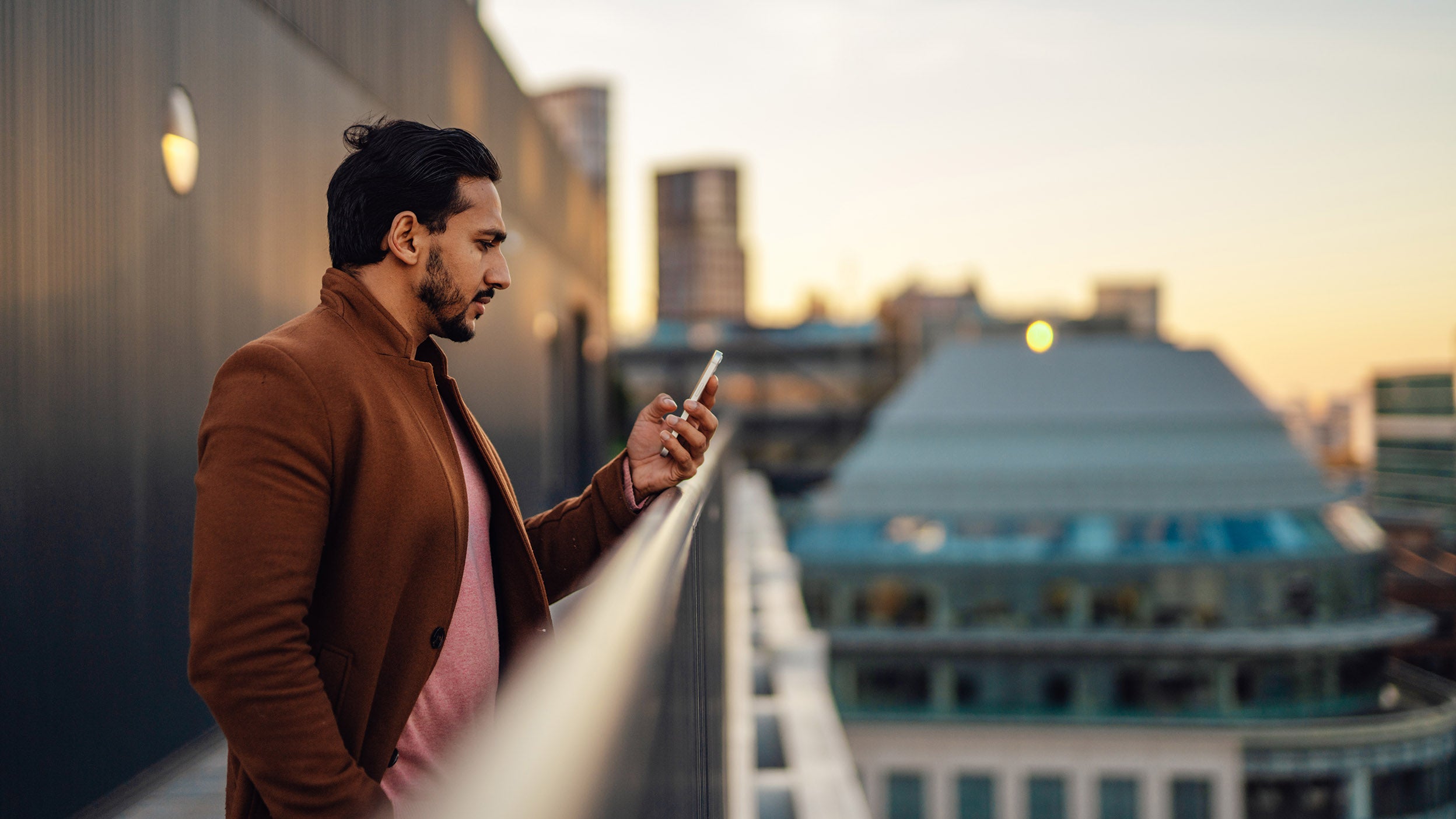 Man stands on a rooftop balcony at sunset, intently looking at his smartphone with city buildings blurred in the background.