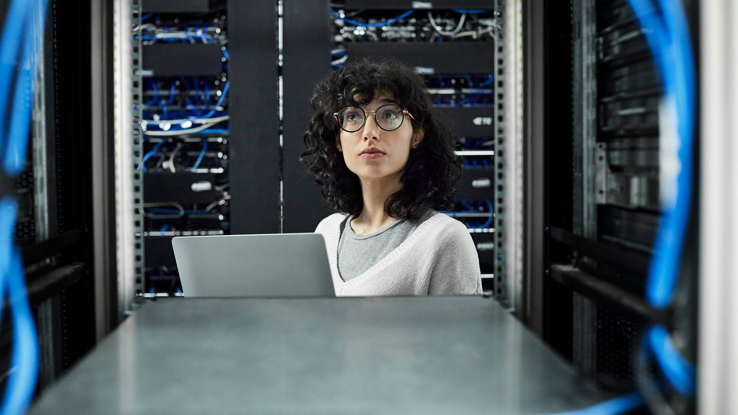 A women with curly hair and glasses looks at her computer while in a data center. Holdings within Invesco QQQ ETF are helping data center efficiency and optimizing AI.
