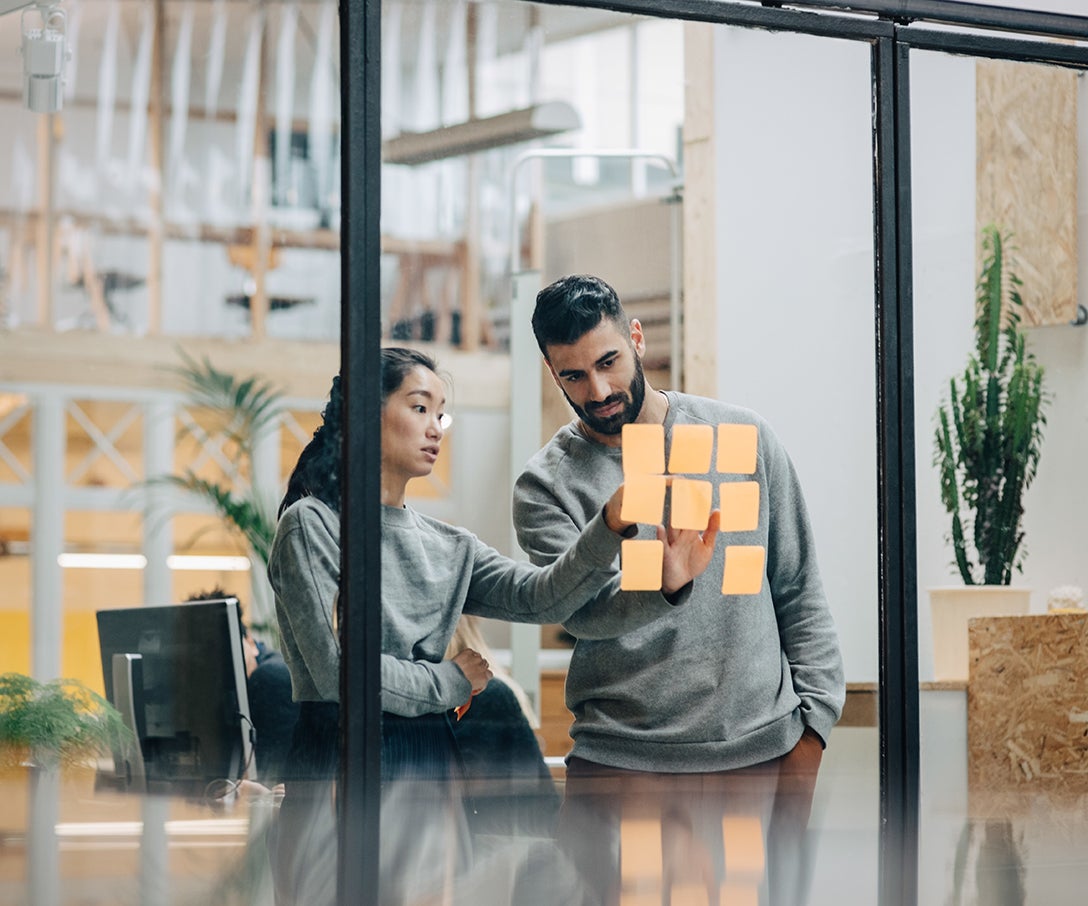 Business persons with sticky notes on glass wall in office discussing research and development topics as innovated by Invesco QQQ holding companies.