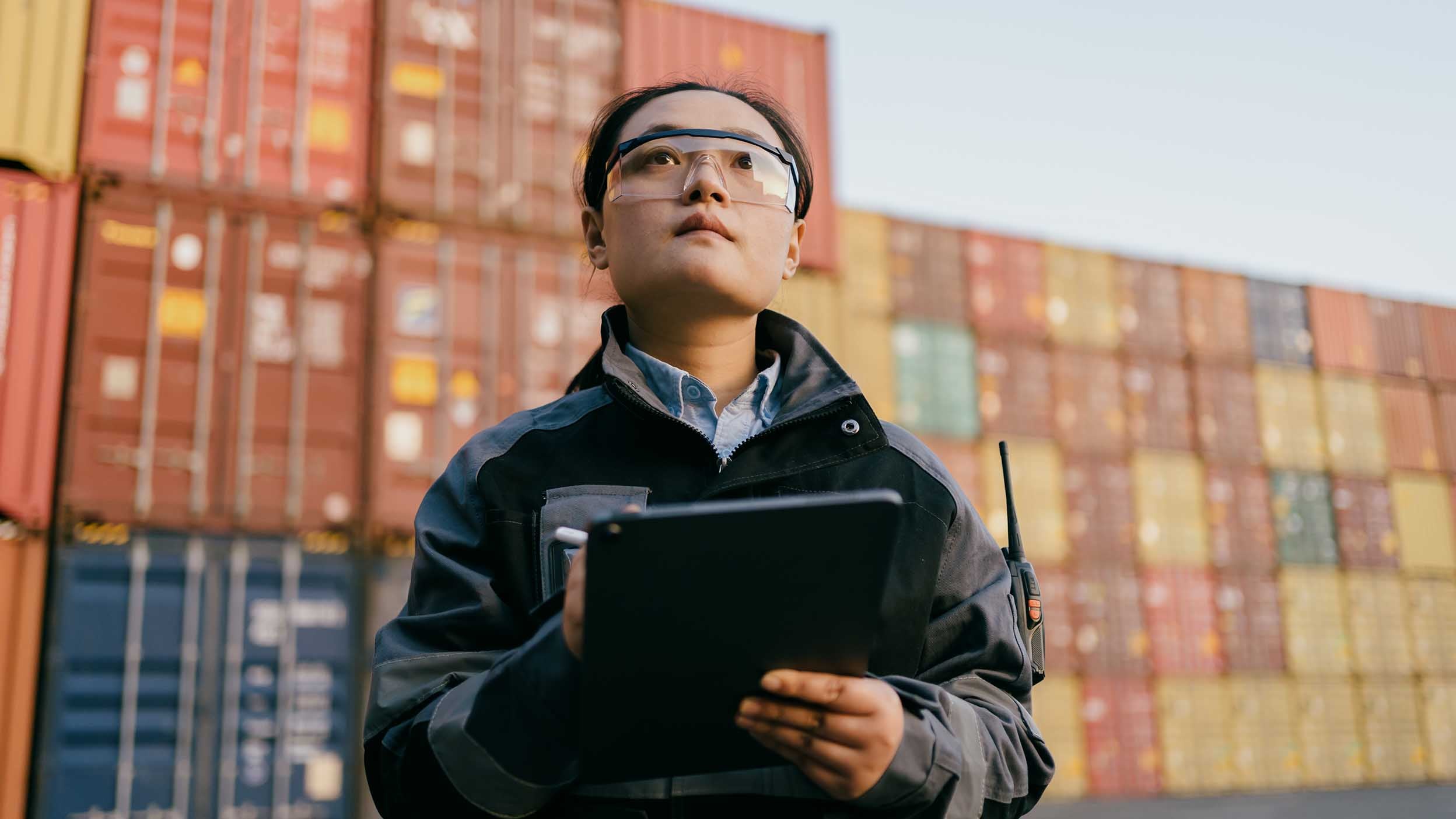 Female Asian inspector wearing safety glasses and carrying a tablet examines containers in a port.