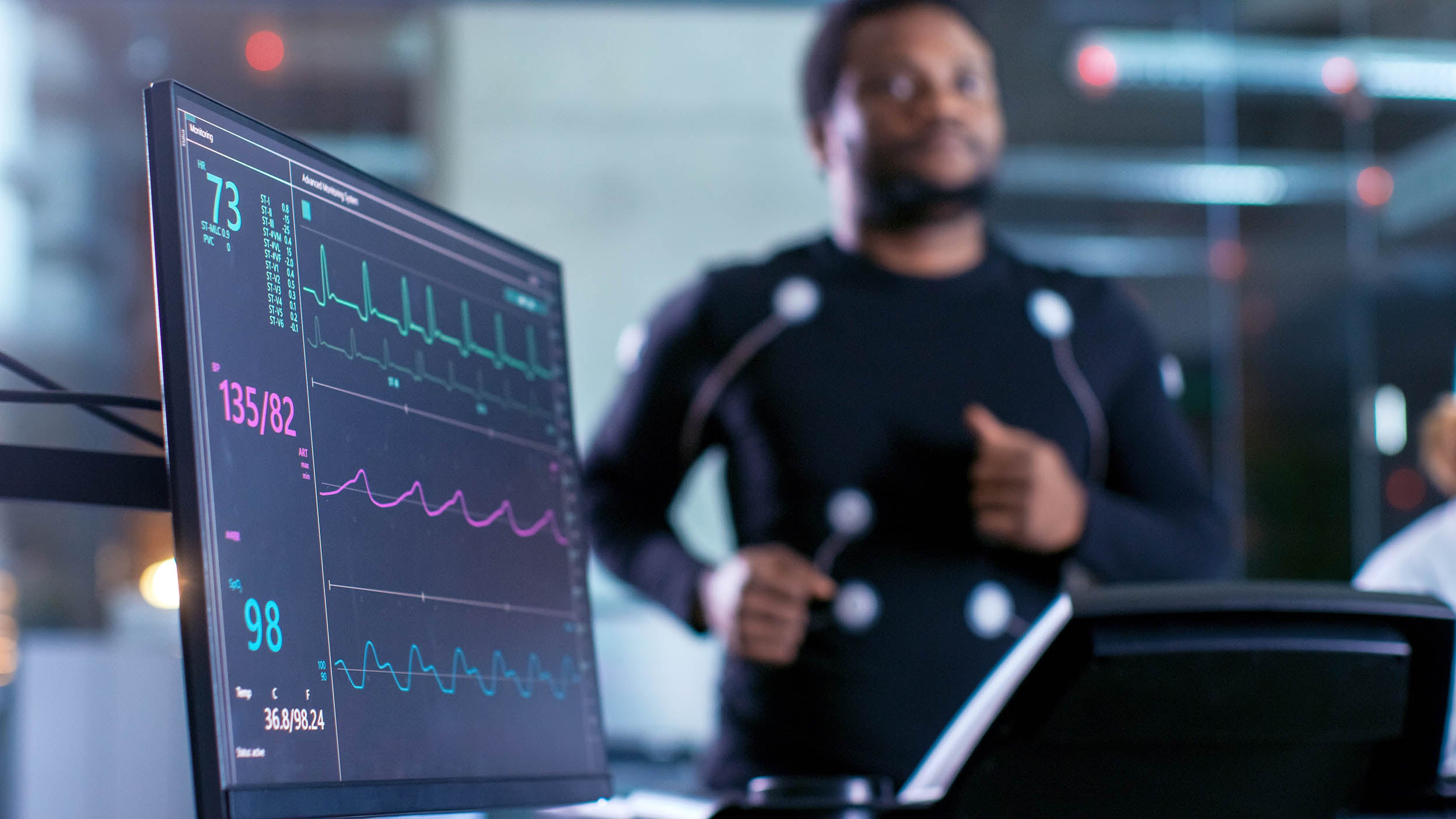 Close-up Shot of a Monitor with EKG Data. Male Athlete Runs on a Treadmill with Electrodes Attached to His Body while Sport Scientist Holds Tablet and Supervises EKG Status in the Background.
