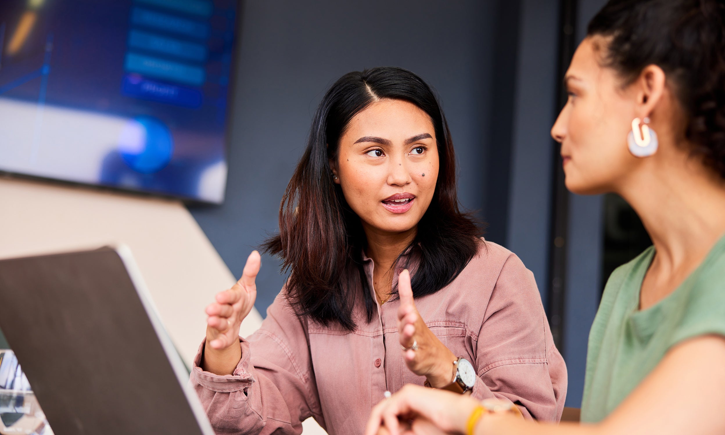 Woman explaining with hand gestures to another woman working on a computer.