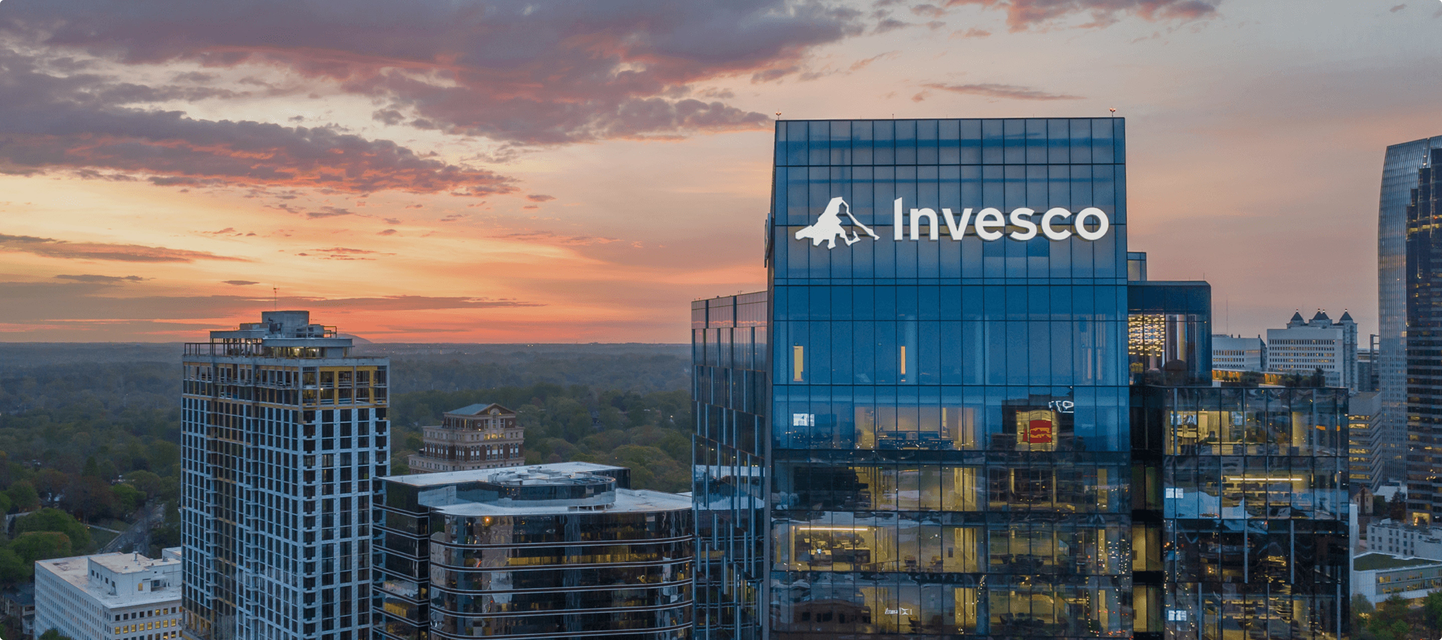 A tall glass office building with the Invesco logo on top, set against a cityscape at sunset with a partly cloudy sky.