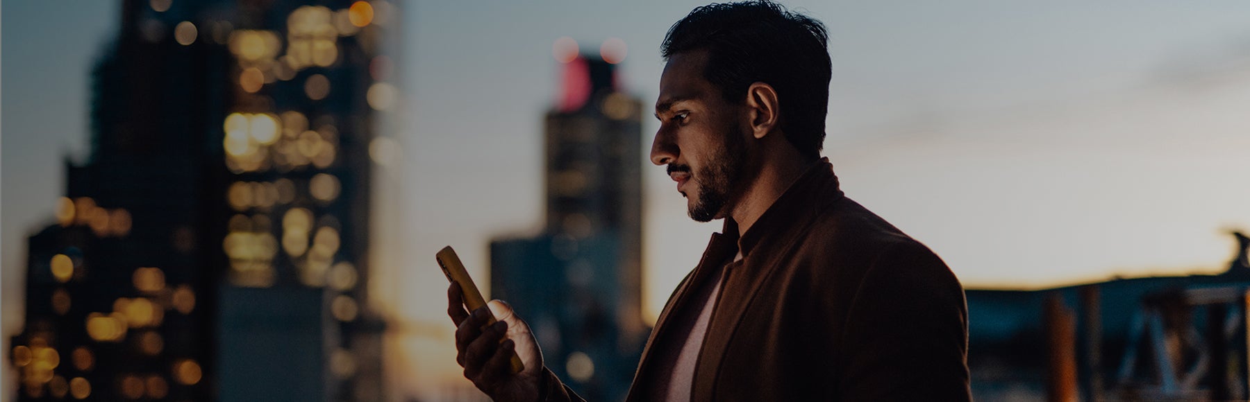 A man stands outdoors in a city at dusk, looking at a smartphone, with tall buildings and lights visible in the background. 