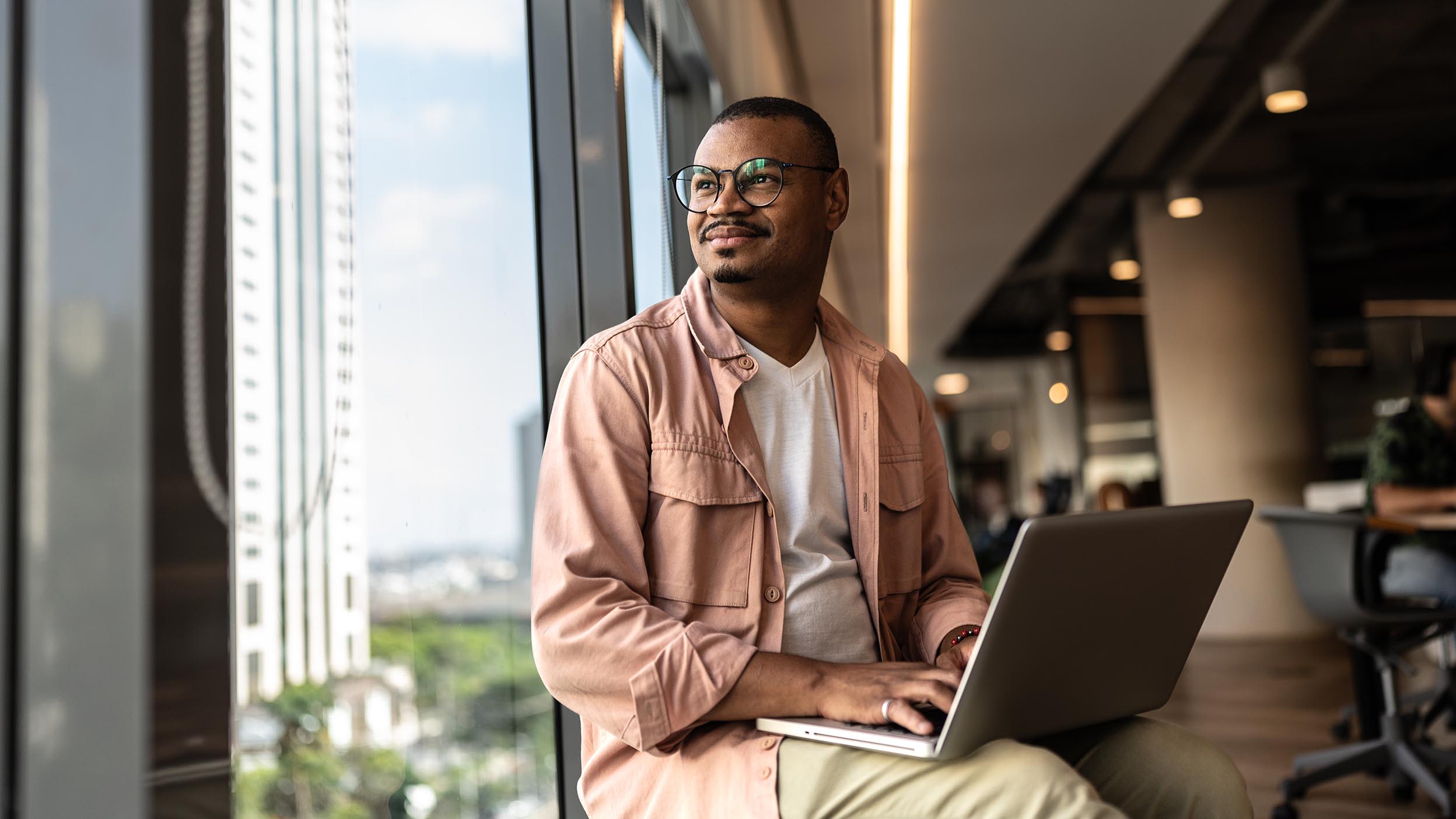 Person by window with laptop on lap in modern indoor setting, seated comfortably.
