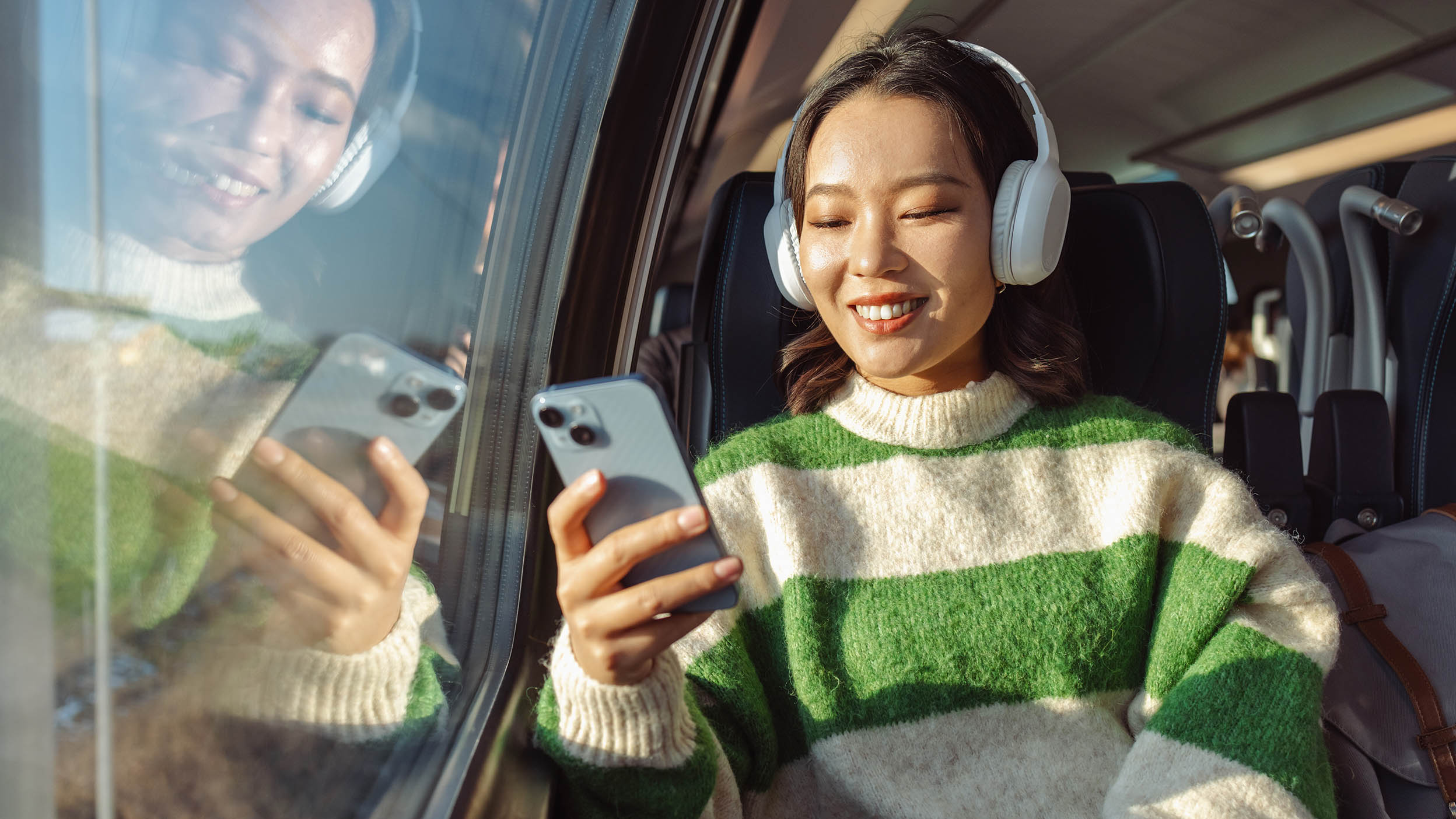 Girl on train wearing headphones, looking at phone, with window and seats in the background.