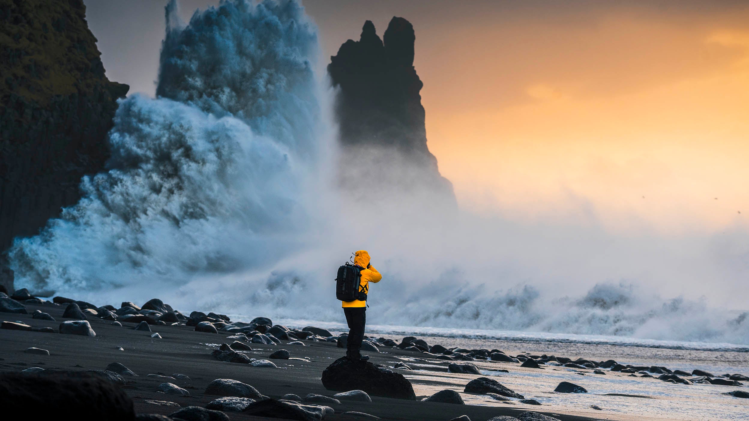 A person in a yellow jacket faces crashing waves and sea stacks at sunset