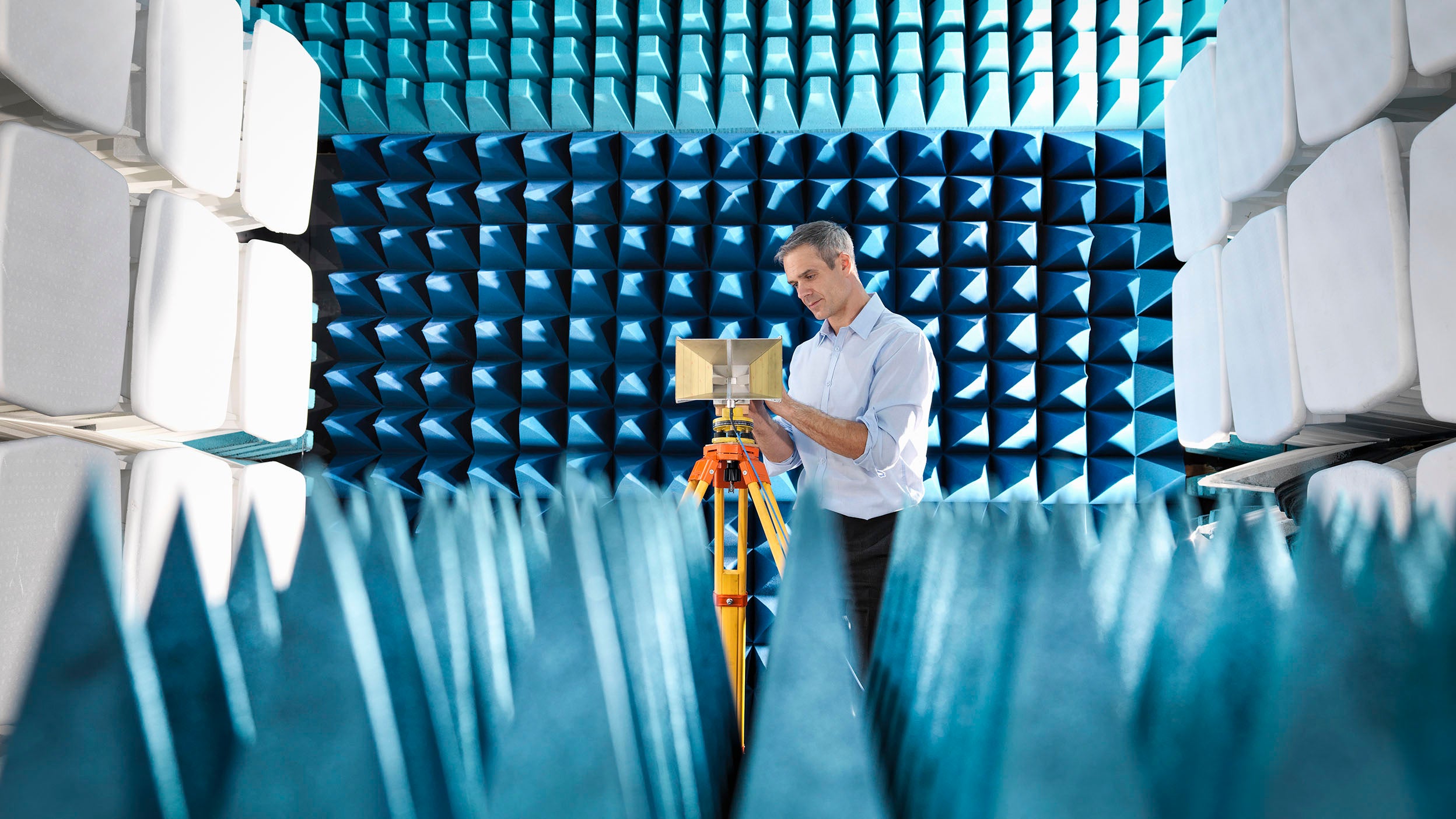 Person in anechoic chamber with blue foam, adjusting tripod-mounted equipment.
