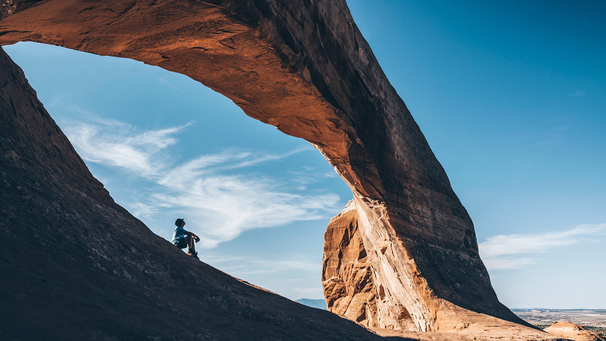 A person sits below a rock arch, looking at the blue sky.